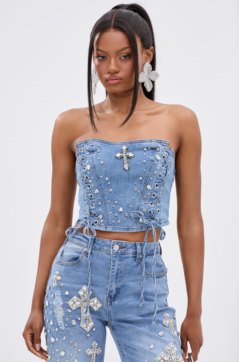 A woman wears the FEELING GRATEFUL RHINESTONE CORSET, a strapless denim corset top with silver studs and a cross, paired with matching high-waisted pants featuring large silver crosses and embellishments. Her long dark hair is styled in a ponytail.