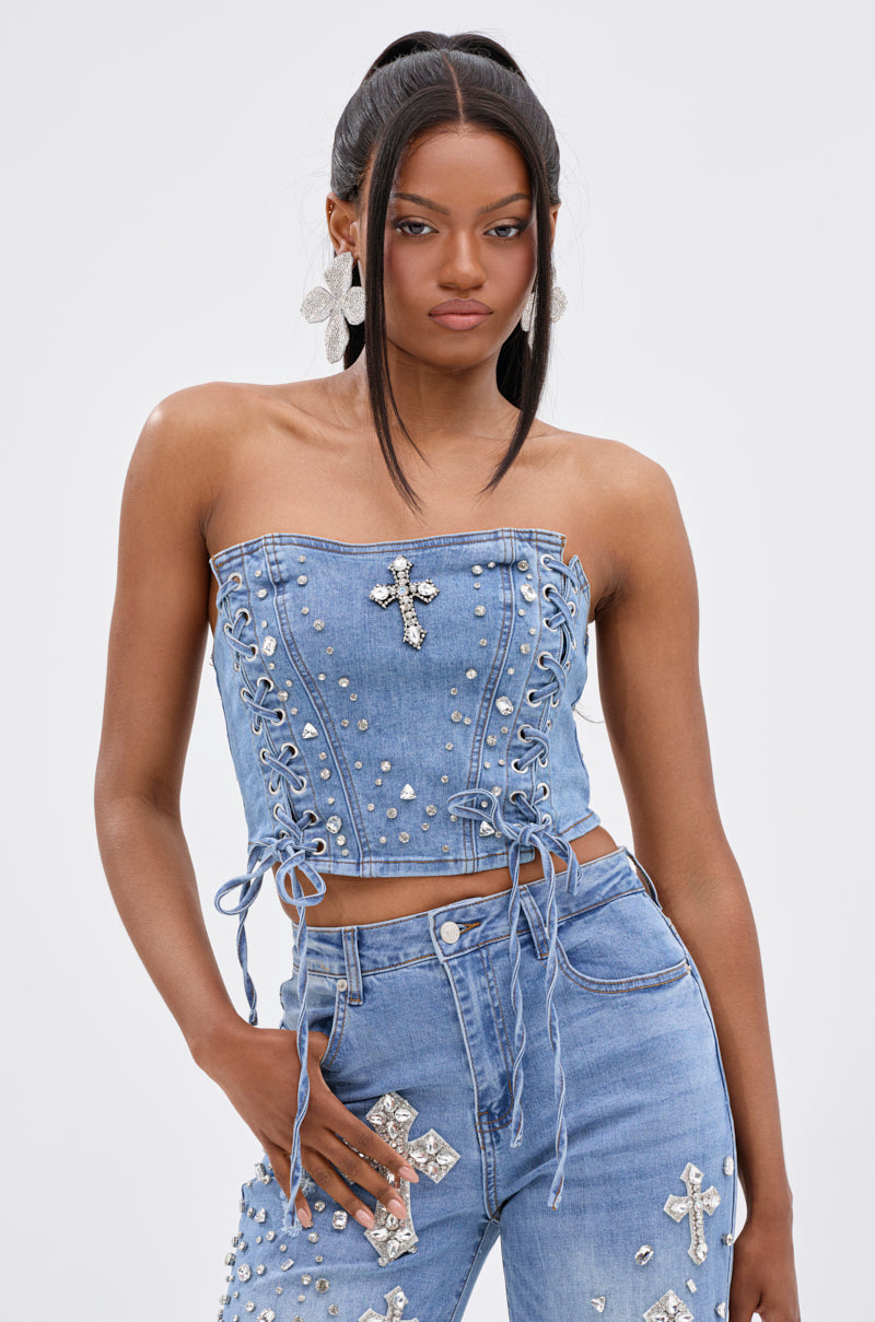 A woman stands confidently against a plain light background, wearing the FEELING GRATEFUL RHINESTONE CORSET—a strapless denim crop top with a lace-up front and rhinestone details—paired with studded, cross-adorned high-waisted jeans.