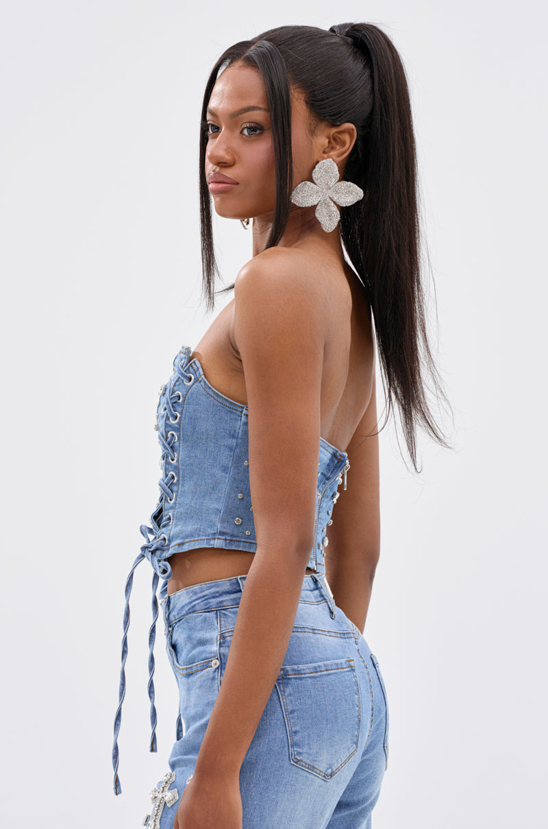 A woman with long straight hair in a high ponytail models the FEELING GRATEFUL RHINESTONE CORSET, paired with matching high-waisted jeans and large silver flower-shaped earrings, standing against a plain light background.