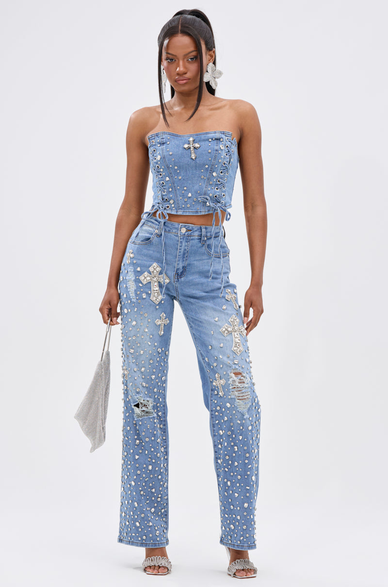 A woman wears the FEELING GRATEFUL RHINESTONE CORSET, a sleeveless denim top with a lace-up front, silver studs, and cross-shaped details, paired with high-waisted jeans. She holds a silver bag and wears heeled sandals on a white background.