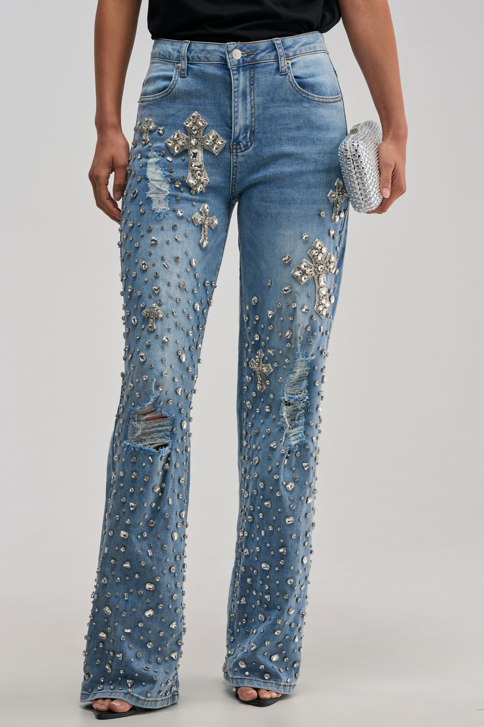Wearing the FEELING GRATEFUL RHINESTONE RELAXED DENIM—a pair of light blue distressed jeans adorned with silver studs and cross-shaped rhinestone accents that channel statement streetwear—this person holds a silver clutch. The upper body is mostly out of frame against a plain light background.