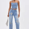 A woman wears the FEELING GRATEFUL RHINESTONE RELAXED DENIM—high-rise jeans with metallic studs and cross motifs—paired with a strapless, lace-up denim top, a silver bag, and heeled sandals.