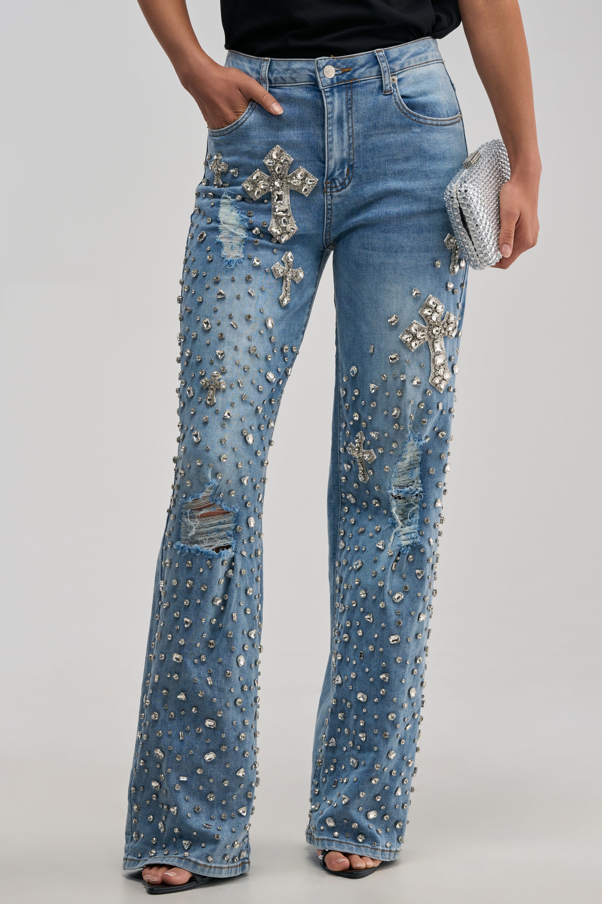 Model is wearing the FEELING GRATEFUL RHINESTONE RELAXED DENIM: light blue, straight-leg, distressed jeans adorned with silver studs and jeweled crosses for a bold streetwear statement, paired with open-toe shoes and accessorized with a sparkly silver clutch bag.