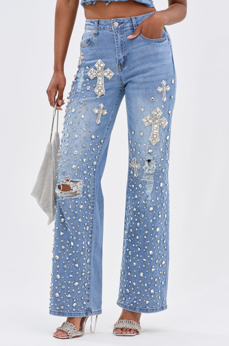 A person wearing FEELING GRATEFUL RHINESTONE RELAXED DENIM with silver cross embellishments, styled with white heels and a sparkly silver handbag.