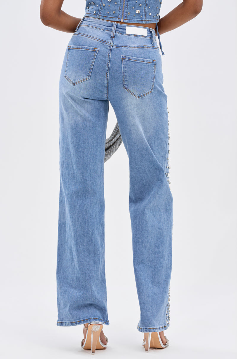 A person shown from the back wears FEELING GRATEFUL RHINESTONE RELAXED DENIM with pearl embellishments on the sides and back pockets, styled with heeled sandals and a studded denim top.