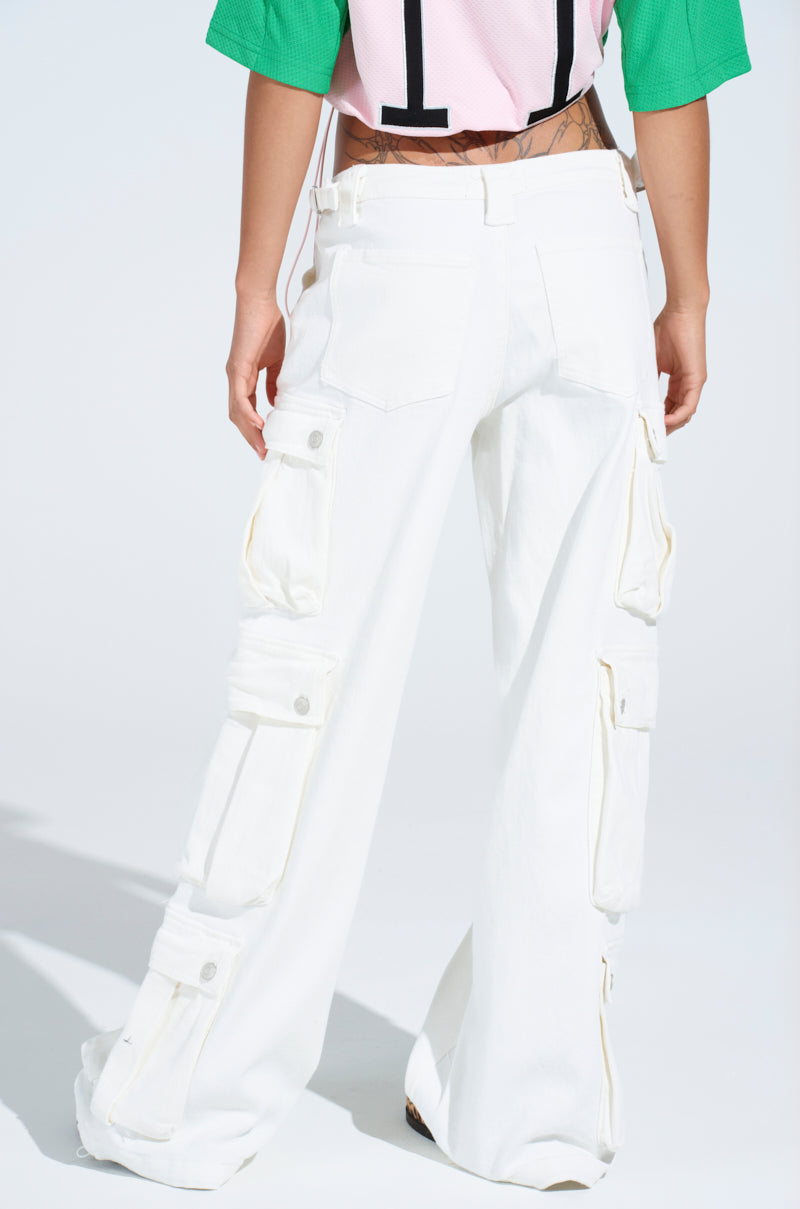 A person wearing "GET RIGHT TO IT CARGO DENIM PANT" in white, featuring multiple pockets and adjustable buckled straps, stands facing away. The pants are paired with a vibrant green and pink top in a well-lit setting against a plain white background.