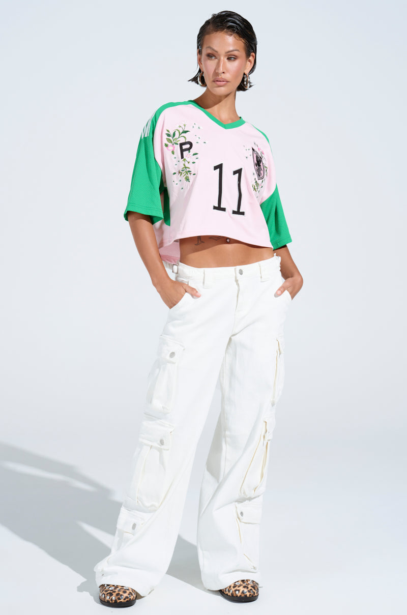 A person poses confidently in a pink and green cropped sports jersey with the number 11, paired with GET RIGHT TO IT CARGO DENIM PANTS featuring adjustable buckled straps and leopard print shoes. The setting is plain and well-lit.
