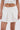 A woman models the ALWAYS GETS BETTER PLEATED MINI SKIRT, a white wrap-style mini with a pleated design, paired with a fitted white corset top. The photo shows her from mid-torso to mid-thigh.