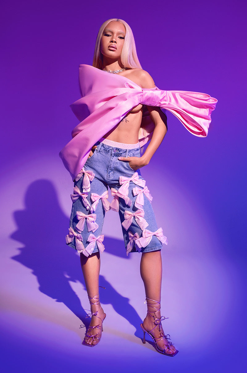 A model with long straight light pink hair and a confident expression wears the BOWS ALL OVER DENIM BERMUDA SHORT, featuring pink organza bows, styled with a pink bow top and strappy pink heels against a purple background.