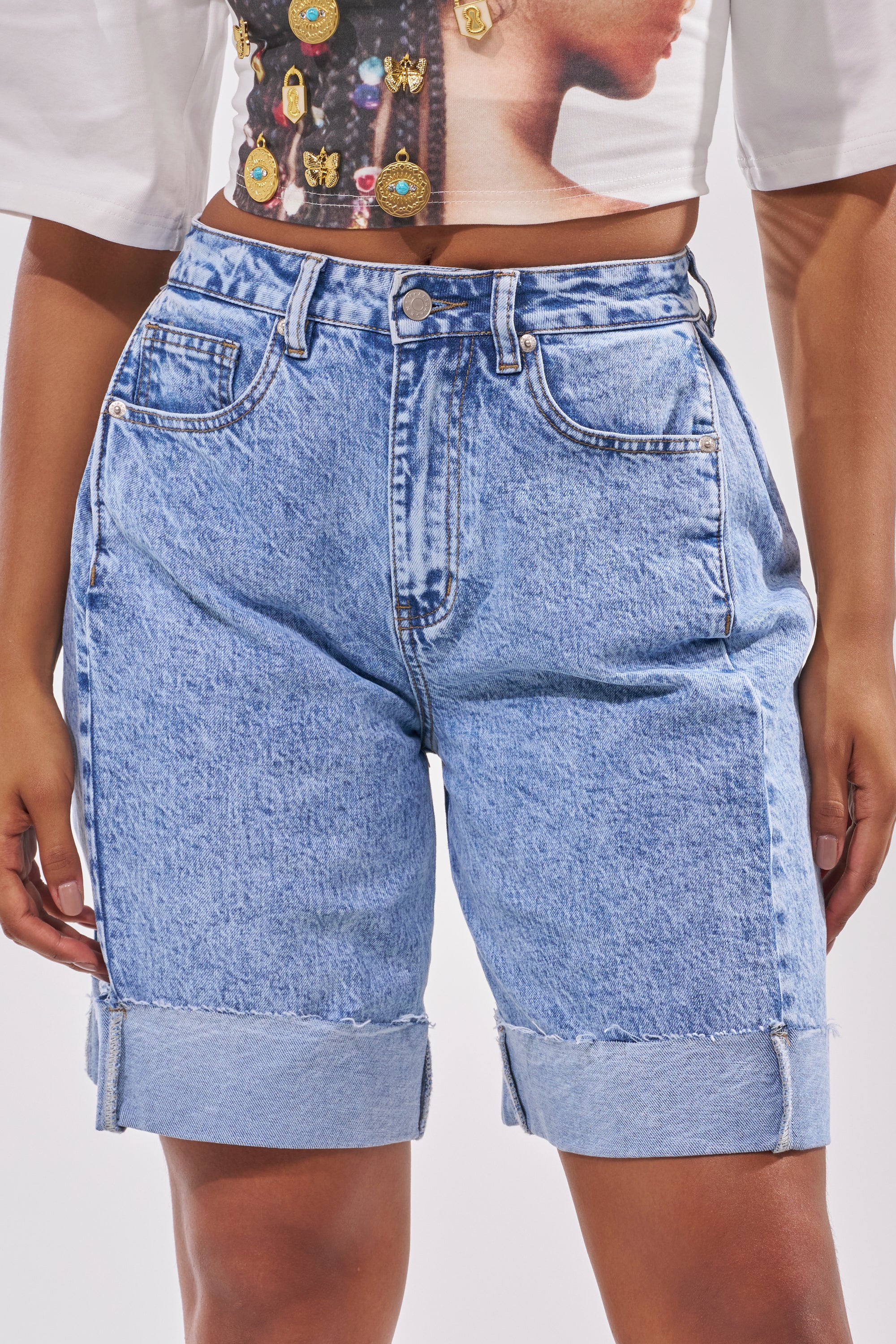 Wearing the GONE BAD DENIM BERMUDA SHORT, a person pairs these high-rise, light-wash shorts with rolled cuffs and a printed crop top showing jewelry and part of a woman's face; only their torso and upper legs are visible. 2