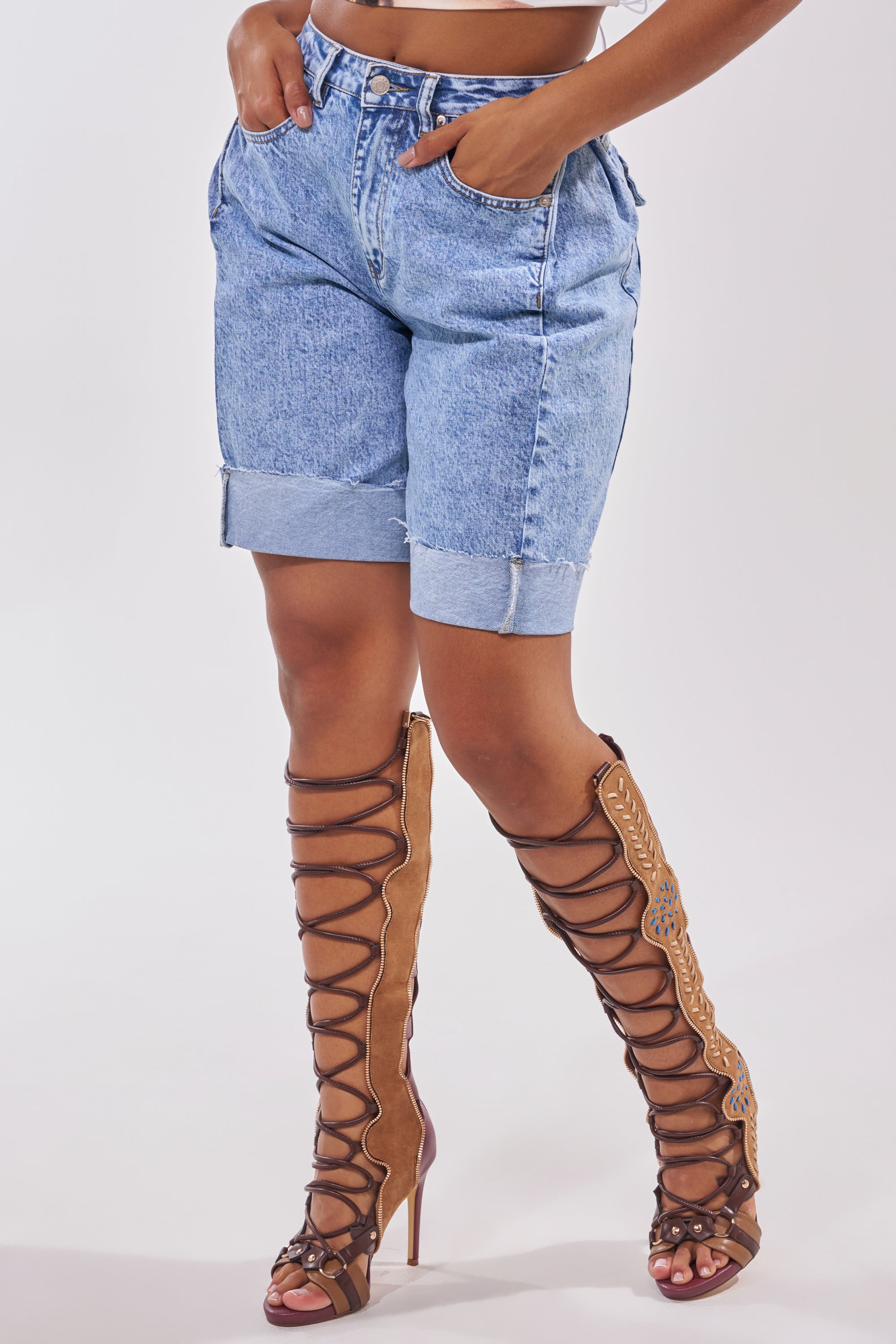 Someone wears the GONE BAD DENIM BERMUDA SHORT in light blue with a cuffed hem, paired with tan, knee-high lace-up high-heeled sandals with cutout details, against a plain white background. 0