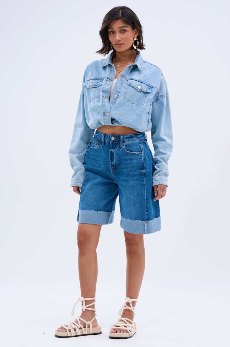 A woman faces the camera in the GONE BAD DENIM BERMUDA SHORT—a high-rise, light blue style with a distressed raw hem—paired with a denim jacket, neutral crop top, white lace-up sandals, hoop earrings, and shoulder-length dark hair.