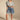 A woman stands confidently in casual attire, wearing a light denim button-up shirt and the GONE BAD DENIM BERMUDA SHORT—high-rise with a distressed raw-hem finish—paired with white lace-up sandals.