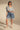 A woman stands confidently in casual attire, wearing a light denim button-up shirt and the GONE BAD DENIM BERMUDA SHORT—high-rise with a distressed raw-hem finish—paired with white lace-up sandals.