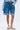 A person wears GONE BAD DENIM BERMUDA SHORTS with a light blue denim shirt and white lace-up sandals, standing against a plain light background.
