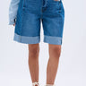 A person wears GONE BAD DENIM BERMUDA SHORTS with a light blue denim shirt and white lace-up sandals, standing against a plain light background.