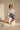 A woman with short dark hair and gold hoop earrings poses confidently in a studio, seated on a white cube. She wears the GONE BAD DENIM BERMUDA SHORT with a distressed raw-hem finish, paired with a light denim jacket, white top, and lace-up sandals.