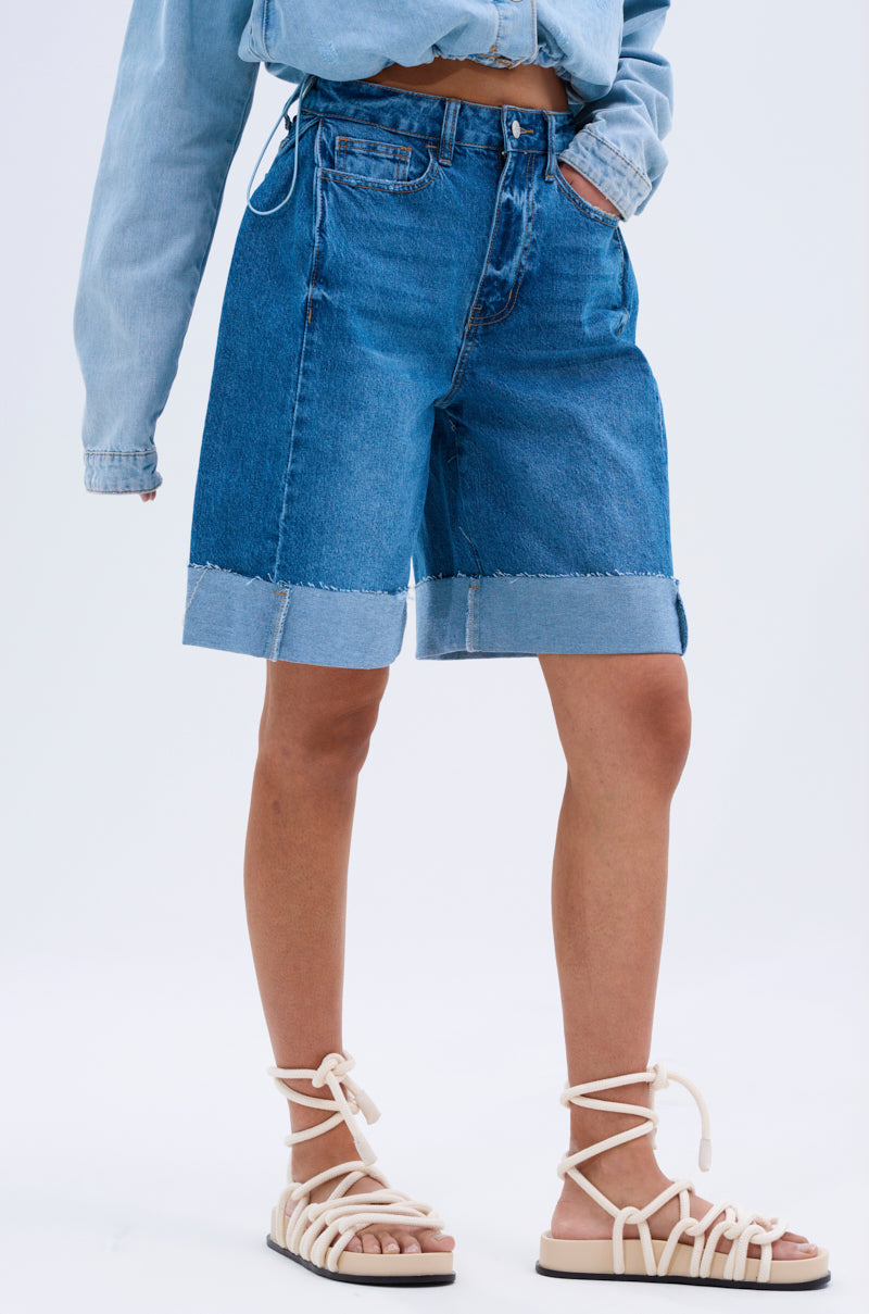 A person in a light blue denim shirt, GONE BAD DENIM BERMUDA SHORTS with a distressed raw-hem finish, and white strappy sandals stands against a plain background.