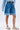 A person in a light blue denim shirt, GONE BAD DENIM BERMUDA SHORTS with a distressed raw-hem finish, and white strappy sandals stands against a plain background.
