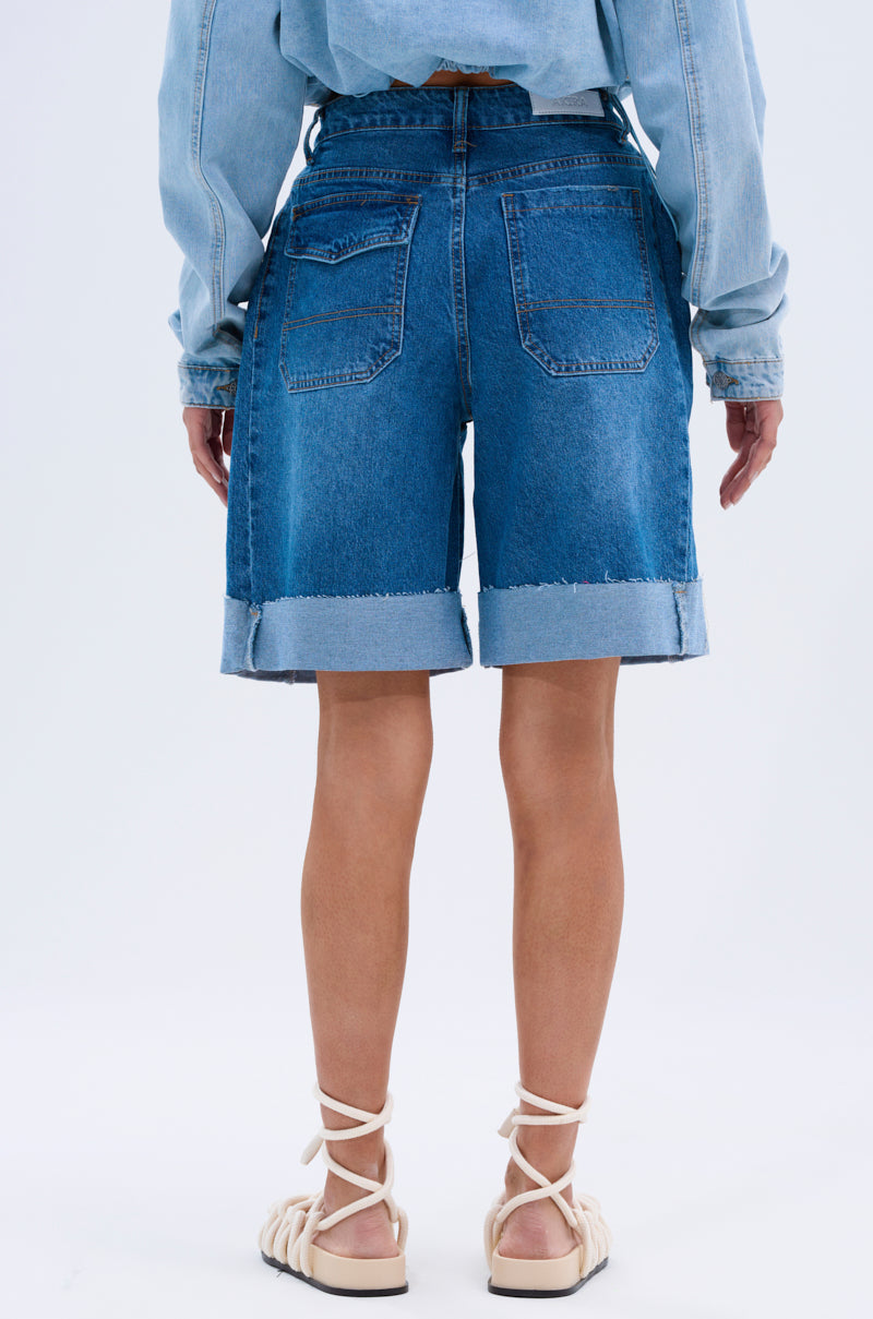 Facing away against a plain white background, a person models the GONE BAD DENIM BERMUDA SHORT with a high-rise fit, distressed raw hem, paired with a light blue denim jacket and white lace-up sandals.