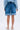 Facing away against a plain white background, a person models the GONE BAD DENIM BERMUDA SHORT with a high-rise fit, distressed raw hem, paired with a light blue denim jacket and white lace-up sandals.