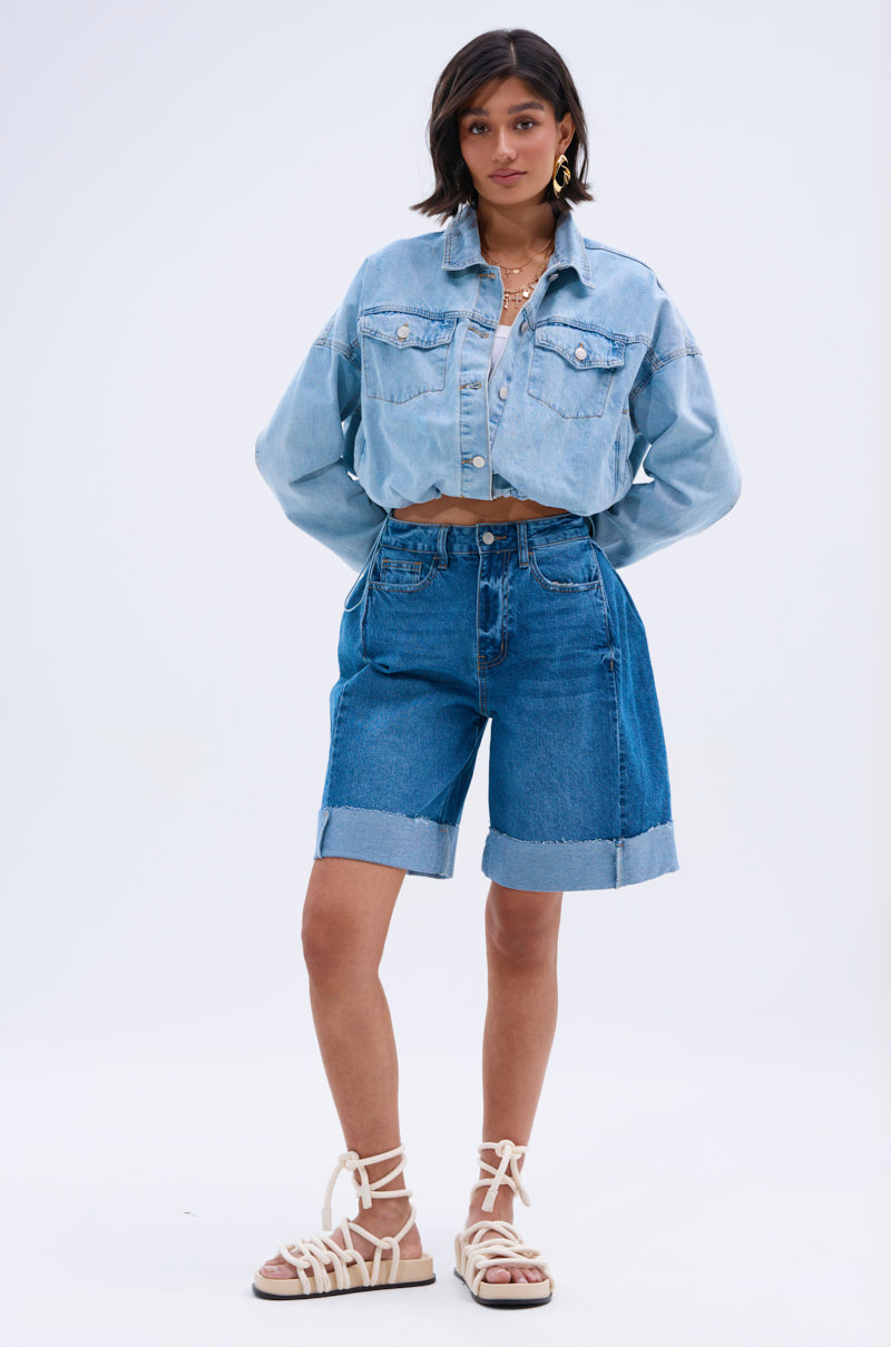 A woman with short dark hair poses in a light blue cropped denim jacket and the GONE BAD DENIM BERMUDA SHORT, featuring a distressed raw-hem finish, paired with white lace-up sandals and layered necklaces against a plain background.