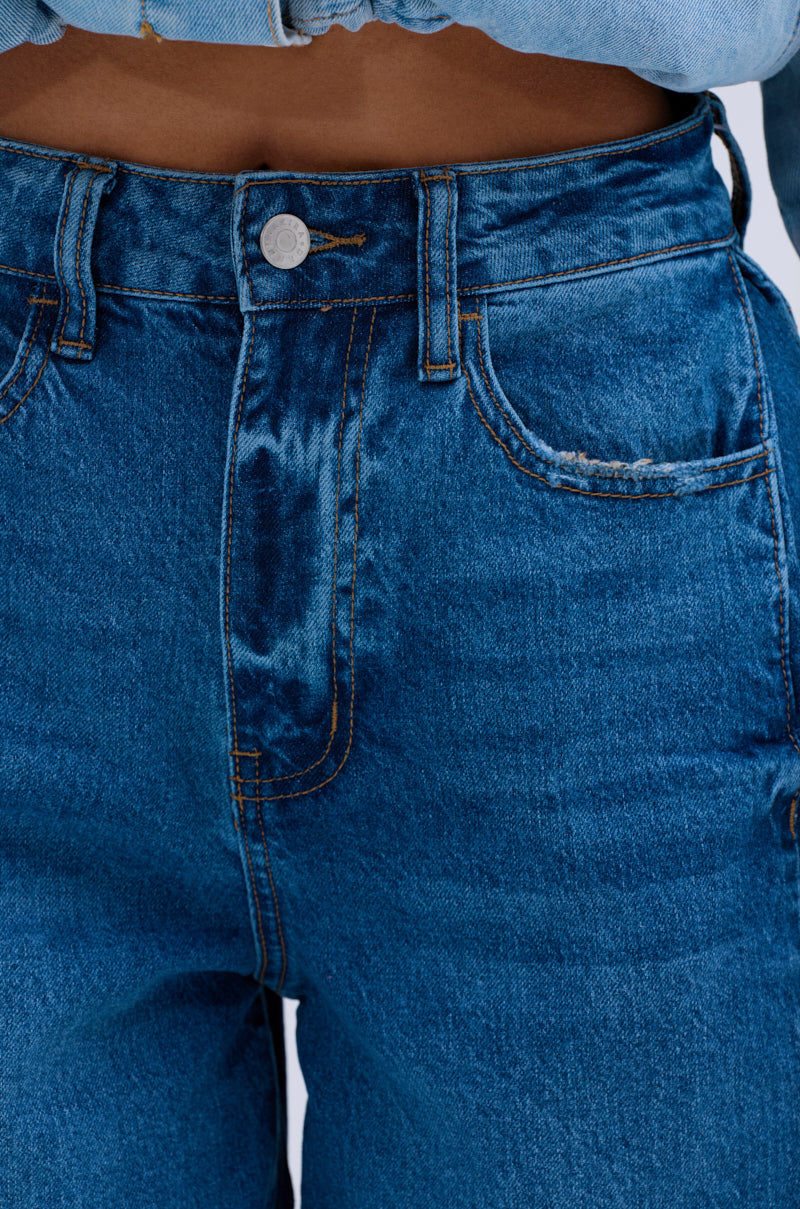 Close-up of a person wearing the GONE BAD DENIM BERMUDA SHORT—high-waisted blue denim with orange stitching, silver button, and distressed raw-hem—paired with a tucked-in light blue denim shirt.