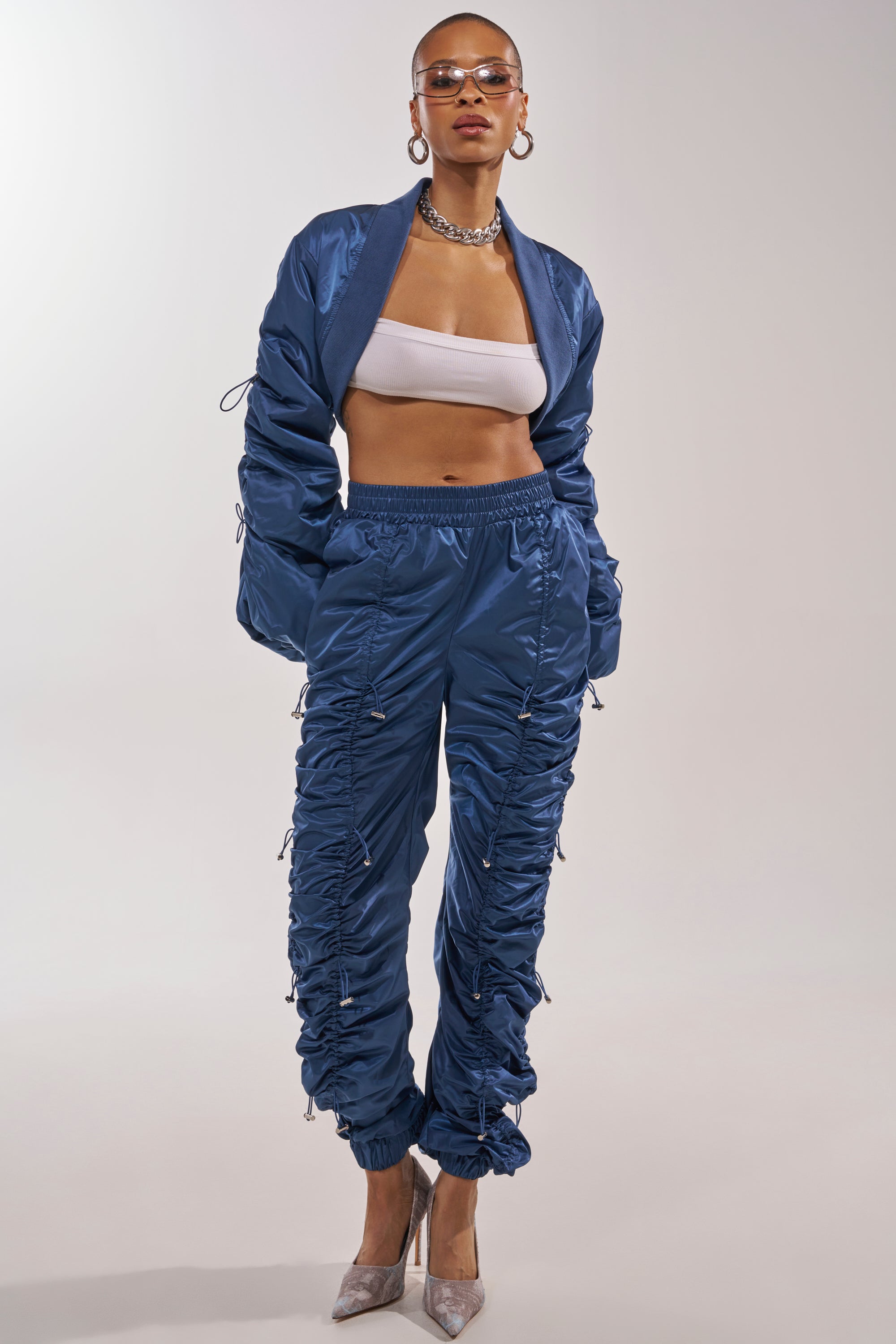 A confident woman in sunglasses wears the NOT TIME FOR GAMES RUCHED JOGGER IN NAVY with a white bandeau top, blue ruched bomber, hoop earrings, a silver necklace, and high heels as she stands against a plain background. 0