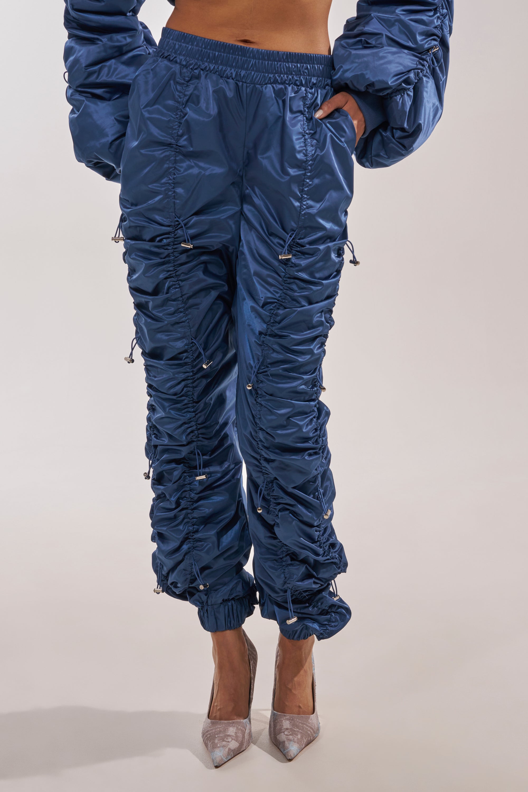 Someone wears the NOT TIME FOR GAMES RUCHED JOGGER IN NAVY featuring shine, ruching, drawstrings, and pockets, paired with pointed-toe silver heels. The upper body is partly shown against a light, plain background. 1