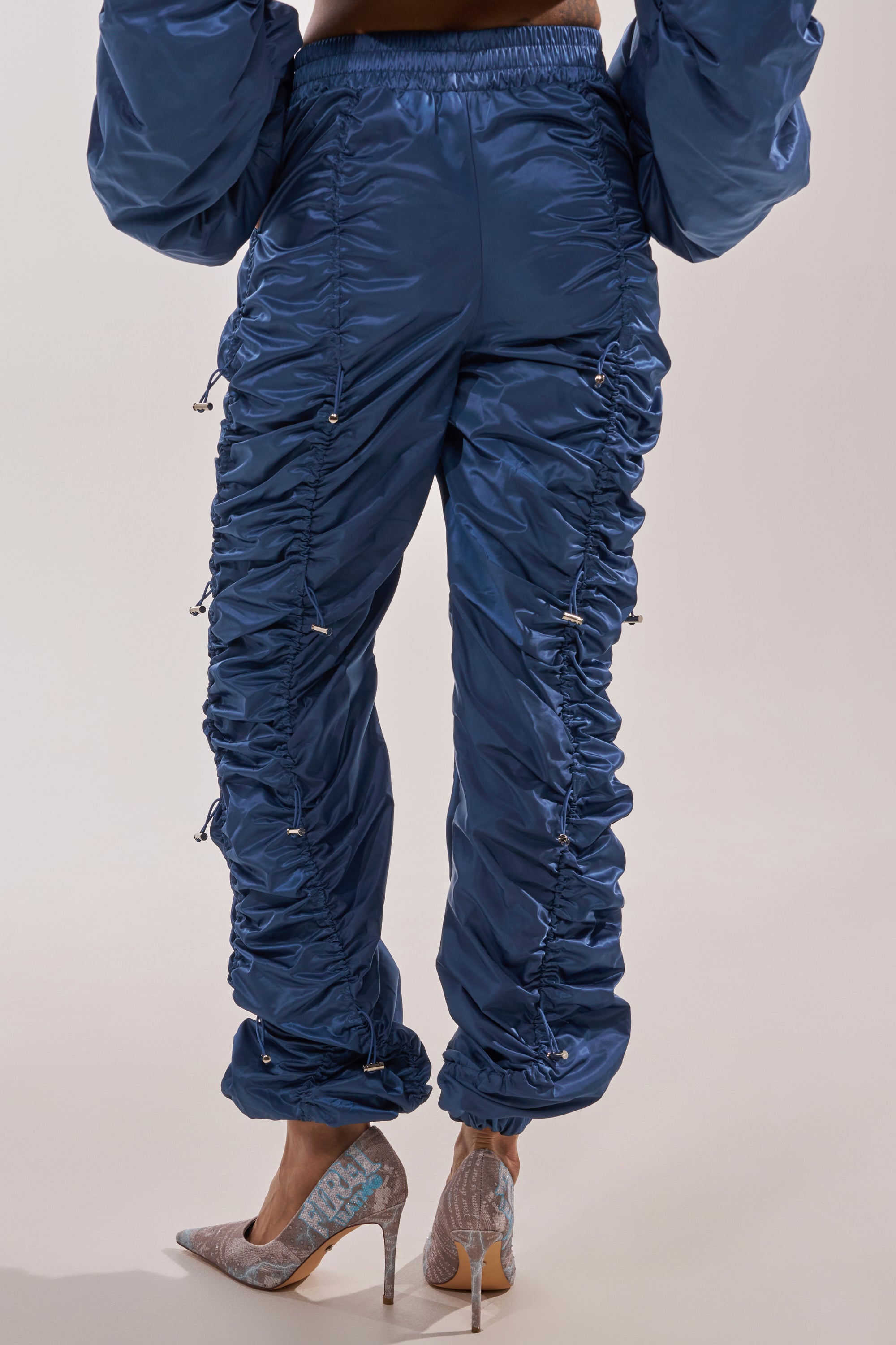 Someone in the NOT TIME FOR GAMES RUCHED JOGGER IN NAVY, featuring shiny blue fabric and adjustable drawstrings, pairs them with patterned high-heeled shoes to highlight streetwear style against a light background. 3