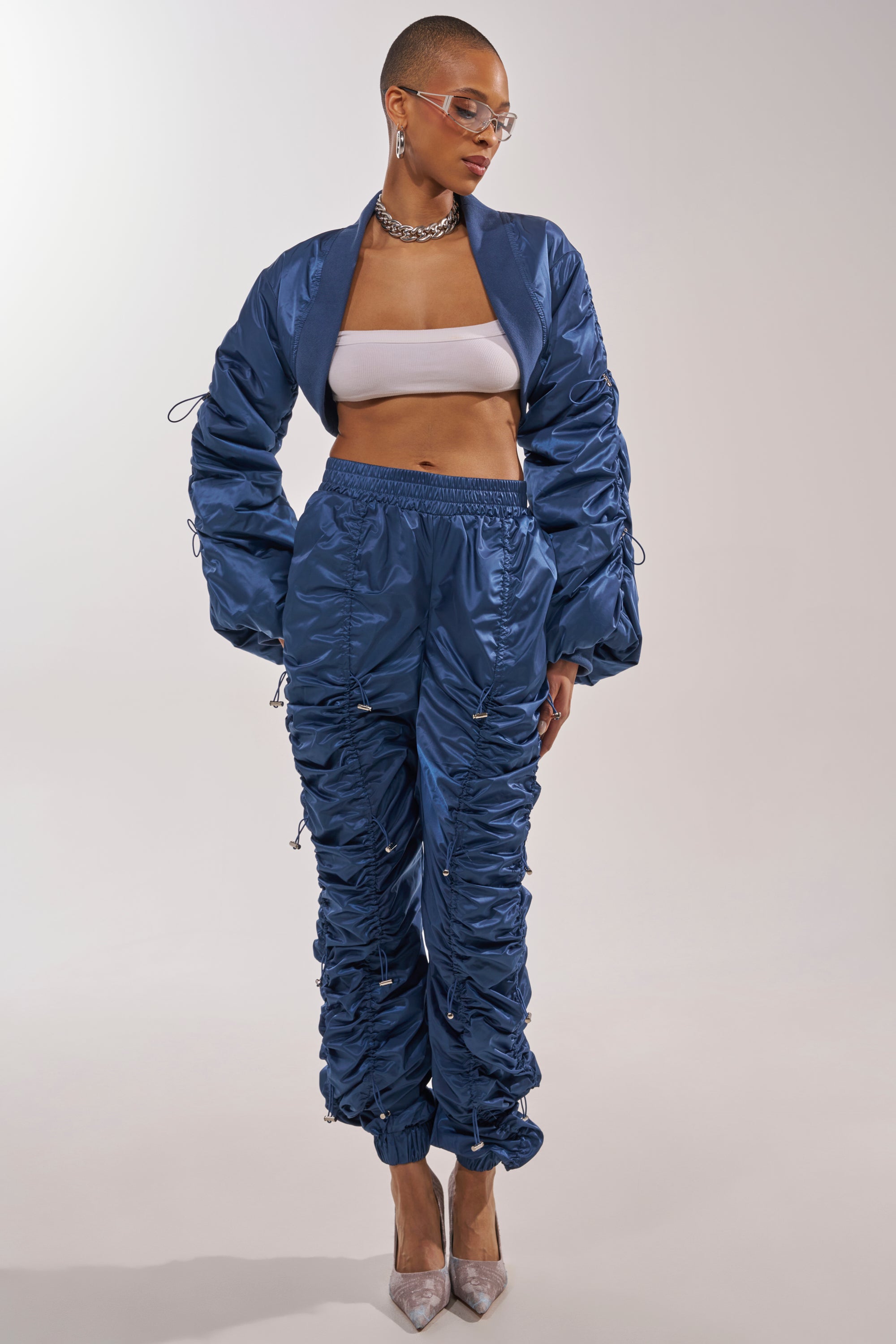 Wearing the NOT TIME FOR GAMES RUCHED JOGGER IN NAVY as part of a blue satin two-piece athleisure set, paired with a white bandeau top, clear glasses, chunky necklace, and silver heels, someone stands against a plain light background. 4