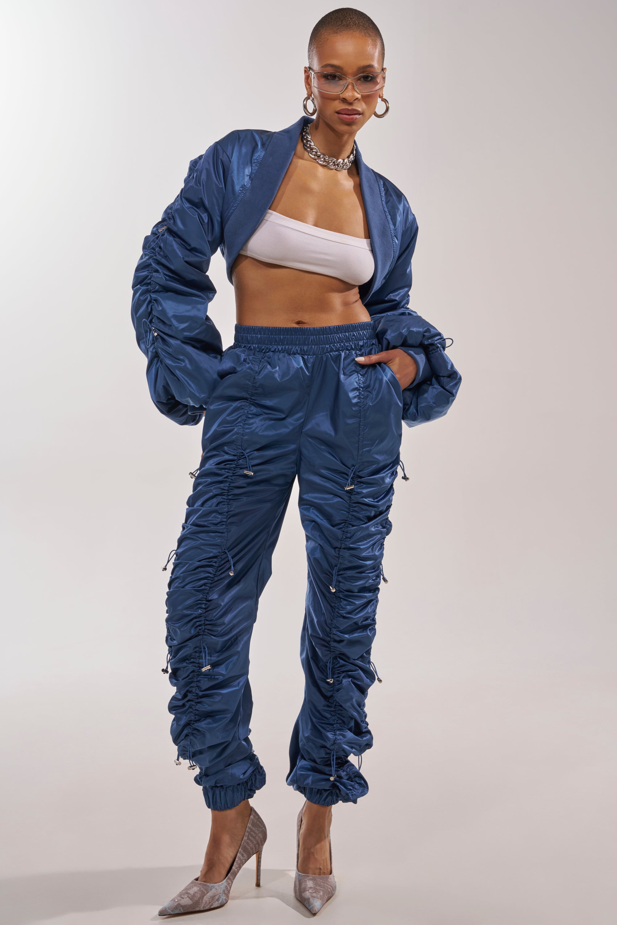 A woman showcases street style in the NOT TIME FOR GAMES RUCHED JOGGER IN NAVY and matching bomber jacket, paired with a white tube top, hoop earrings, sunglasses, chain necklace, and beige pointed heels against a plain backdrop. 5