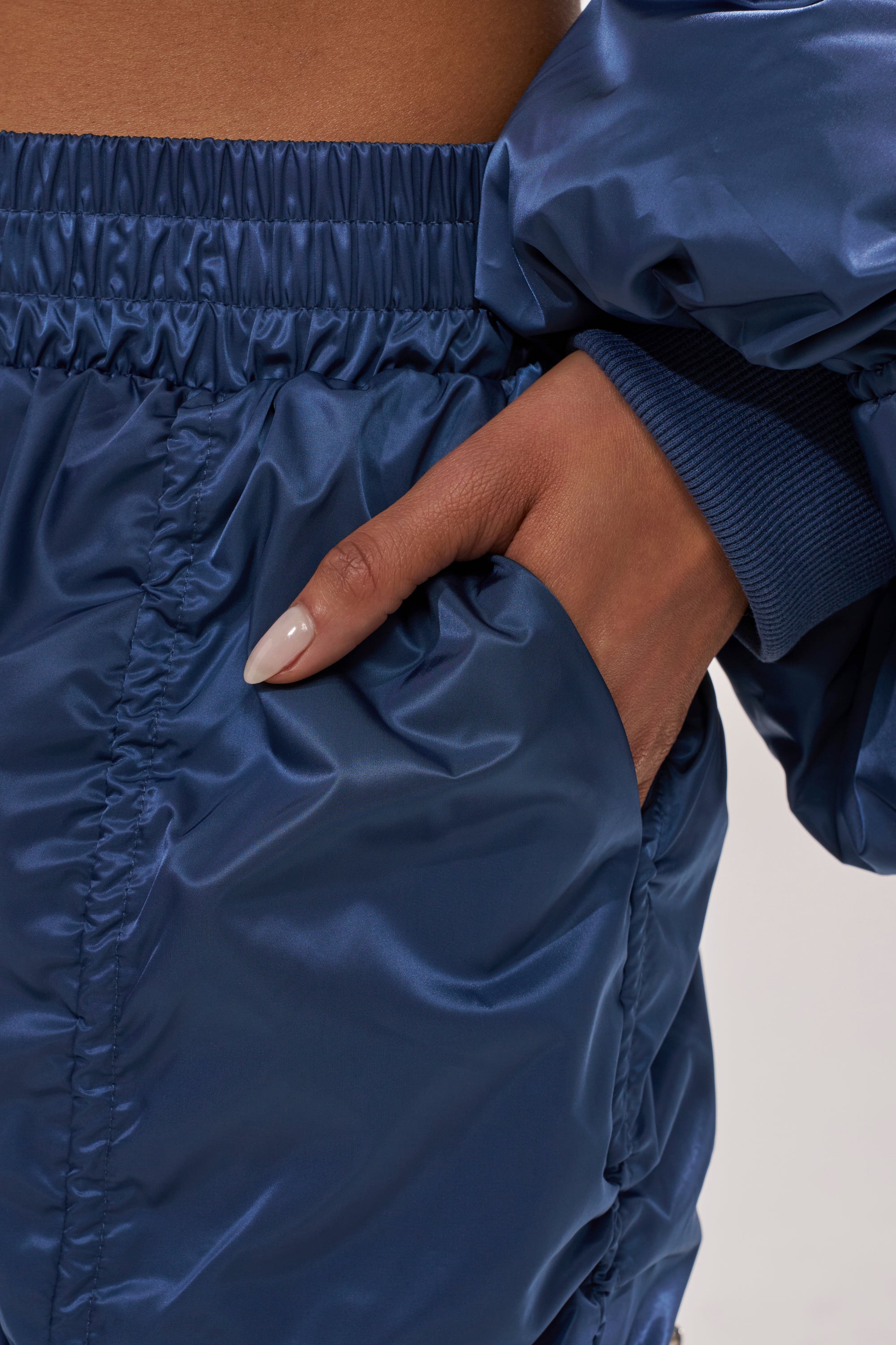 A close-up of a person wearing the NOT TIME FOR GAMES RUCHED JOGGER IN NAVY, featuring shiny blue fabric and a matching jacket, with their hand partially tucked into the jogger pocket. 6