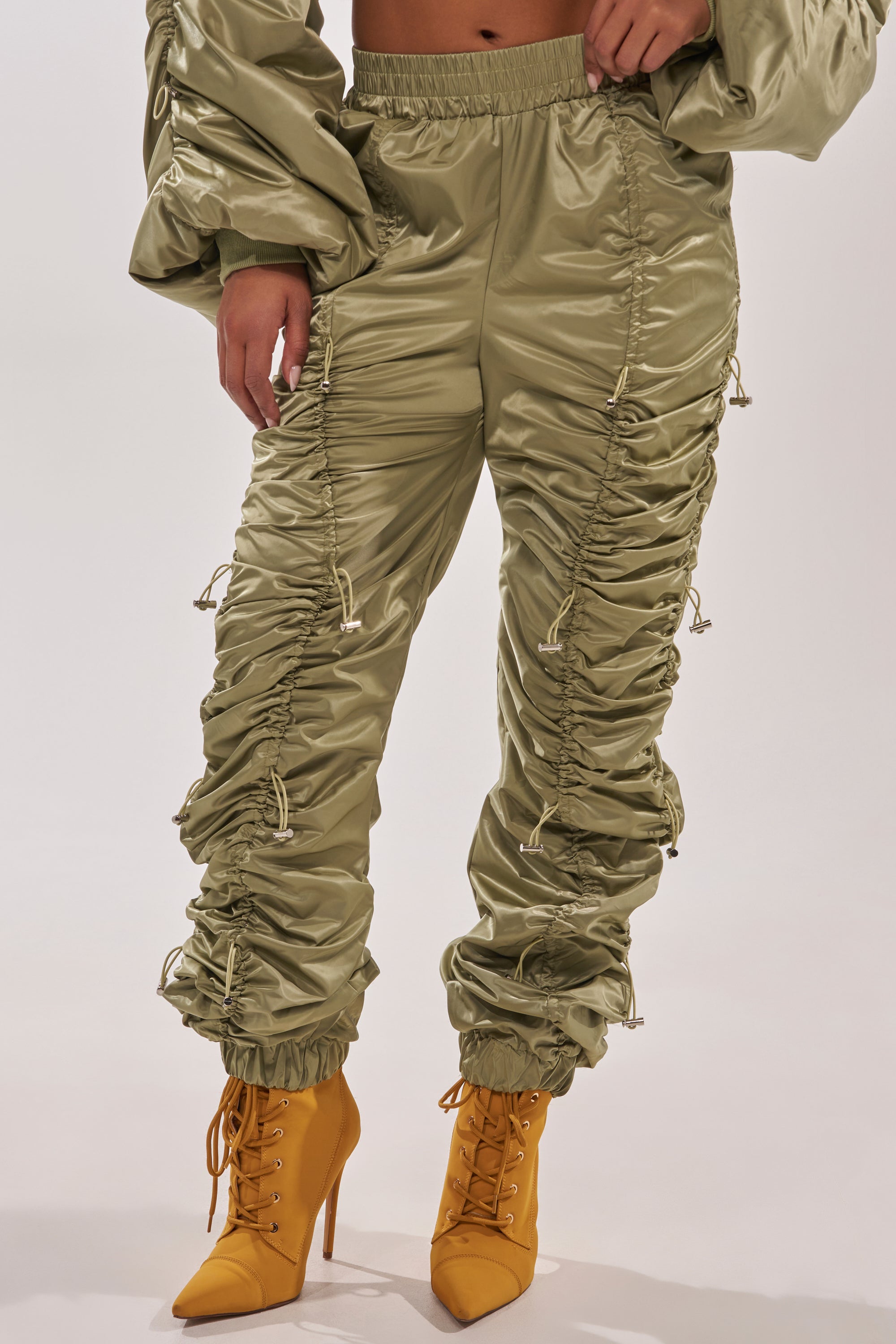 Wearing the NO TIME FOR GAMES RUCHED JOGGER IN OLIVE, featuring a shiny finish and adjustable bungee cord, paired with yellow lace-up high-heeled boots, a person stands against a plain light backdrop. 1