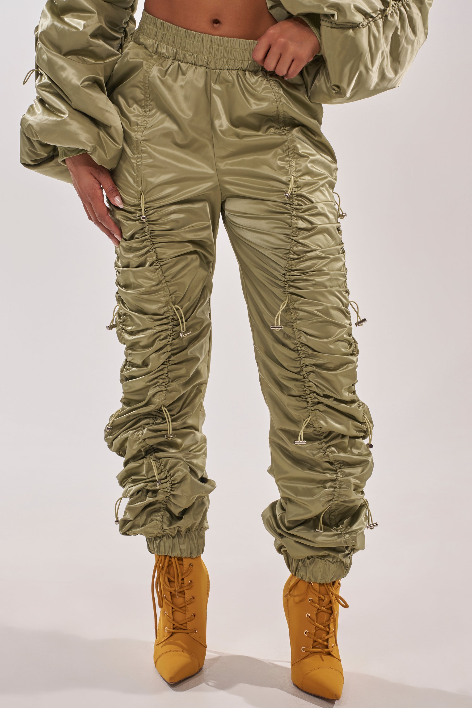 Wearing the NO TIME FOR GAMES RUCHED JOGGER IN OLIVE with drawstrings and puffy sleeves, and paired with mustard yellow lace-up ankle boots, a person stands against a plain light background. 2