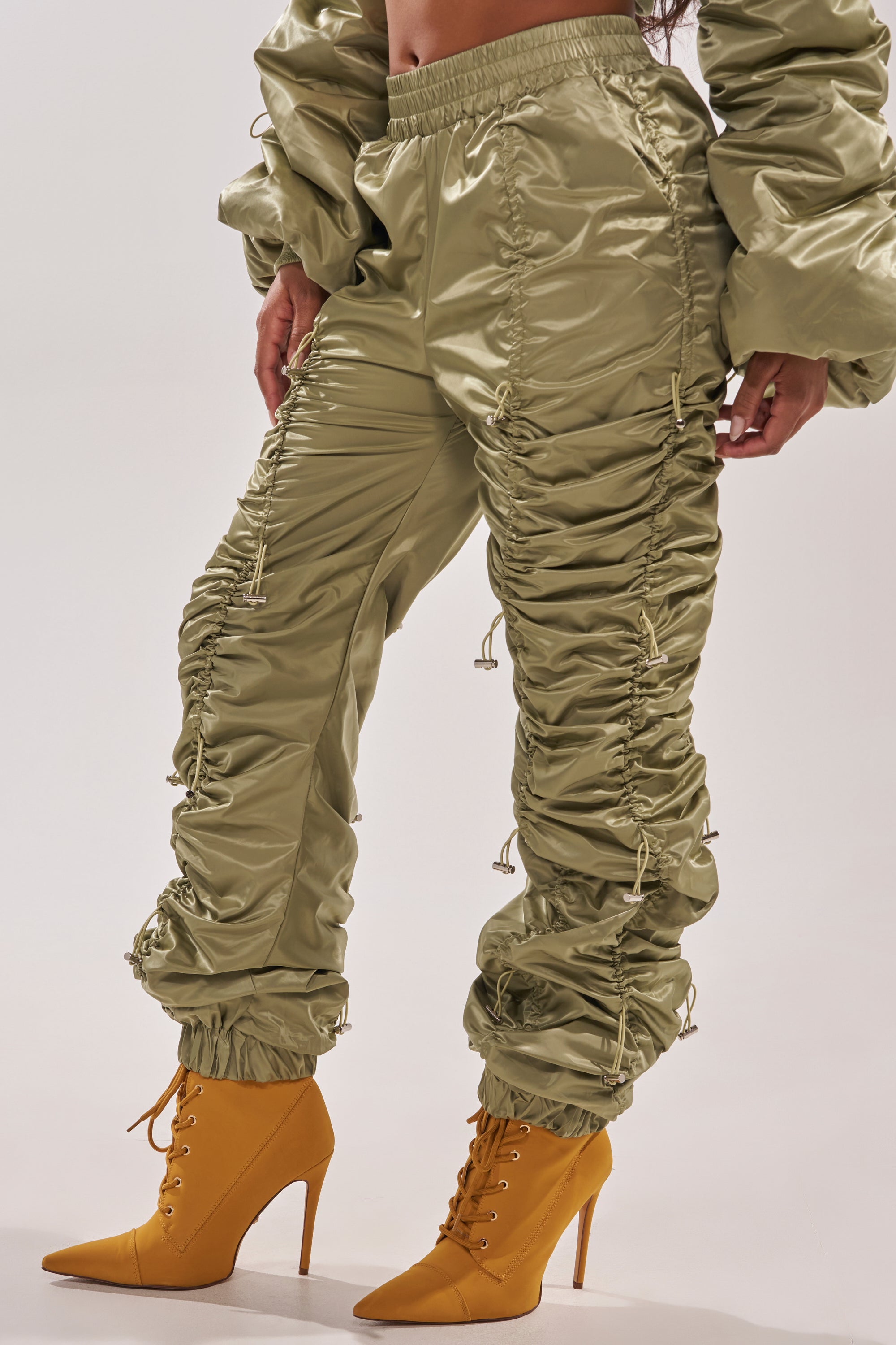 A person wears the NO TIME FOR GAMES RUCHED JOGGER IN OLIVE with adjustable bungee cords and a matching jacket, styled with pointed-toe, lace-up high-heeled mustard yellow boots against a plain white background. 3