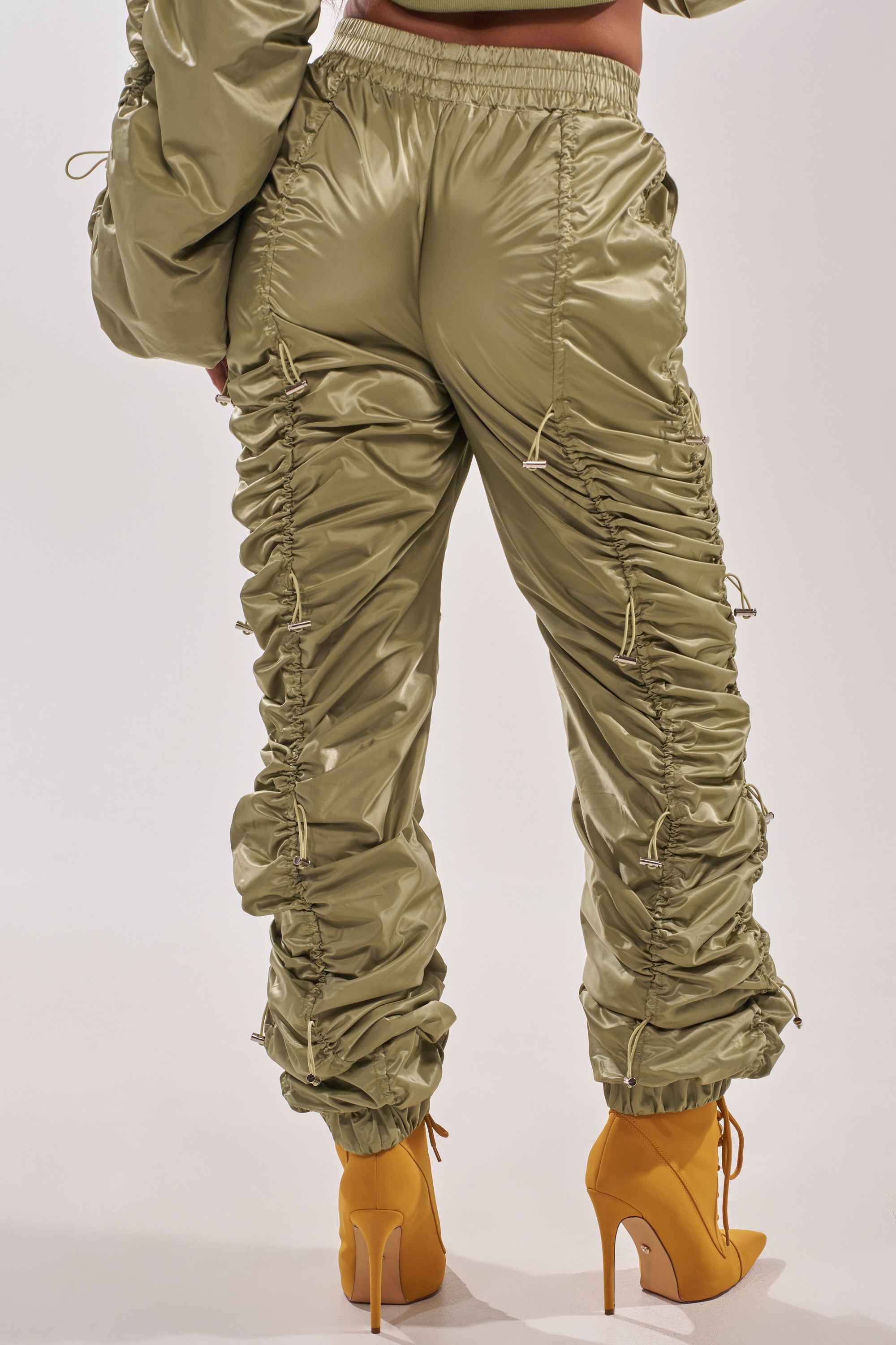 Photographed from the back, a person wears the NO TIME FOR GAMES RUCHED JOGGER IN OLIVE with adjustable bungee cords, styled with mustard yellow high-heeled boots. 4
