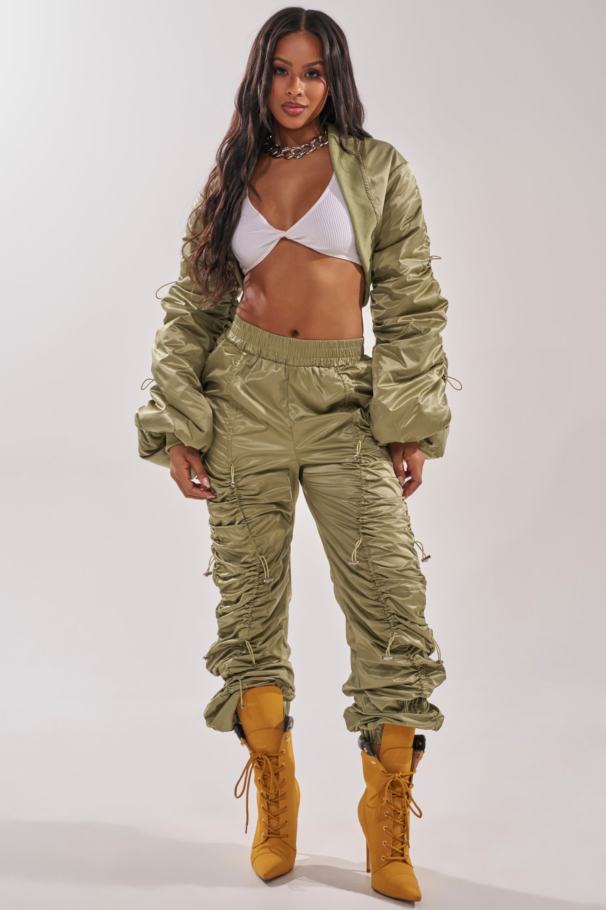 A woman models the NO TIME FOR GAMES RUCHED JOGGER IN OLIVE with a matching ruched jacket, white ribbed crop top, yellow lace-up boots, and a chunky chain necklace against a plain background. 5