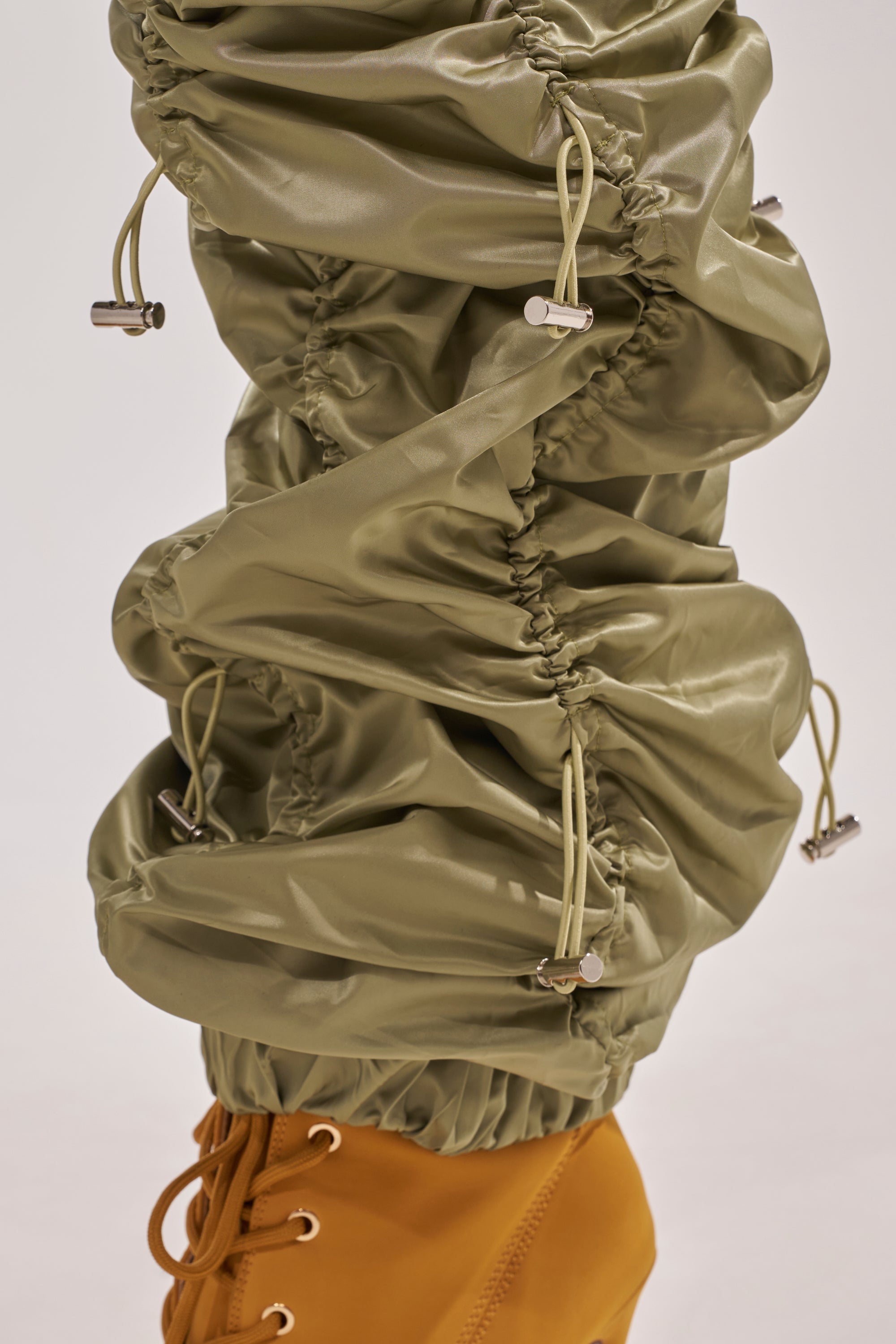 A close-up of the NO TIME FOR GAMES RUCHED JOGGER IN OLIVE, featuring shiny olive-green fabric and an adjustable bungee cord, styled over tan lace-up shoes. 7