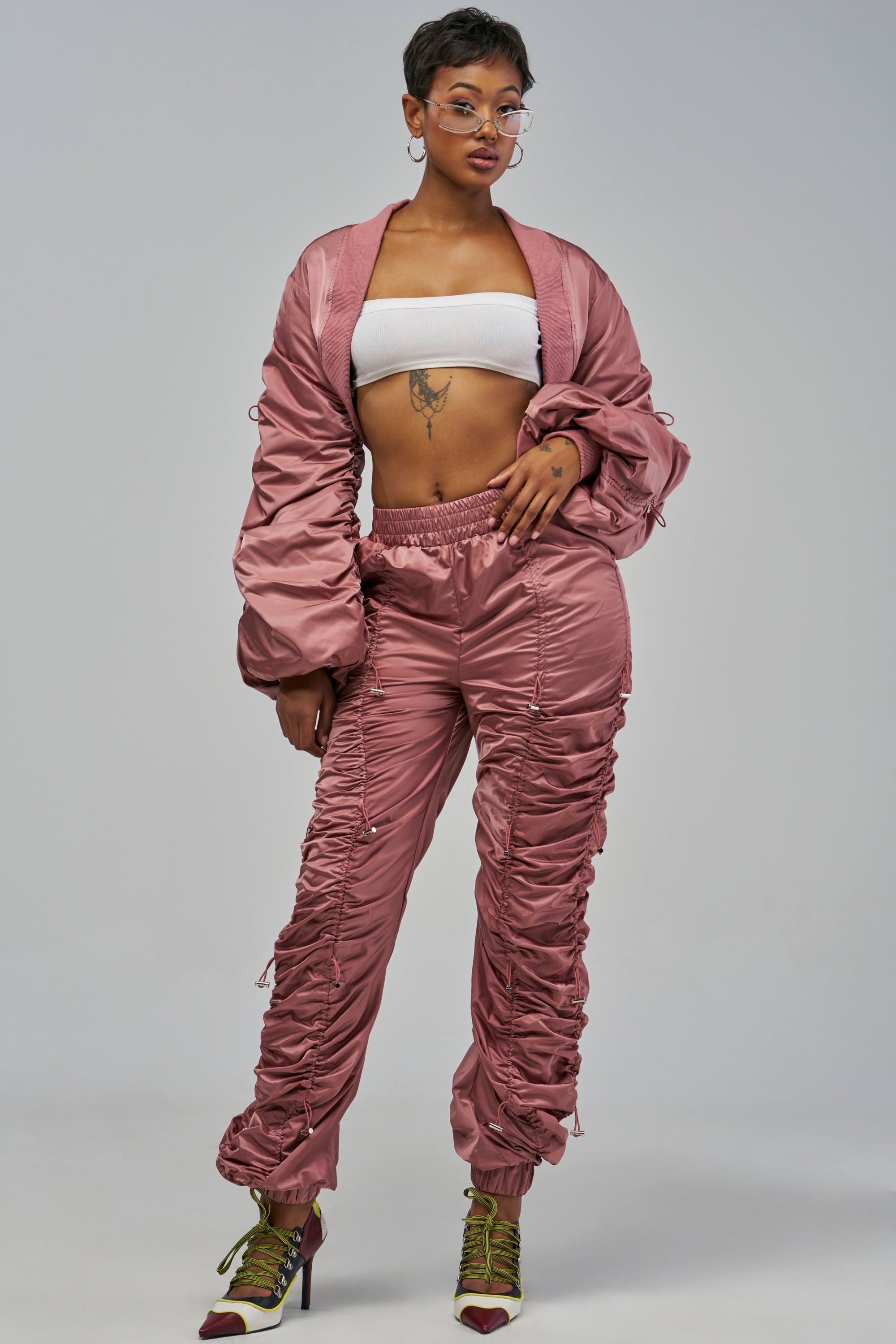 A person wears a mauve NO TIME FOR GAMES RUCHED JOGGER set featuring high-rise pants and a cropped jacket, paired with a white tube top and heeled booties for a stylish streetwear look.