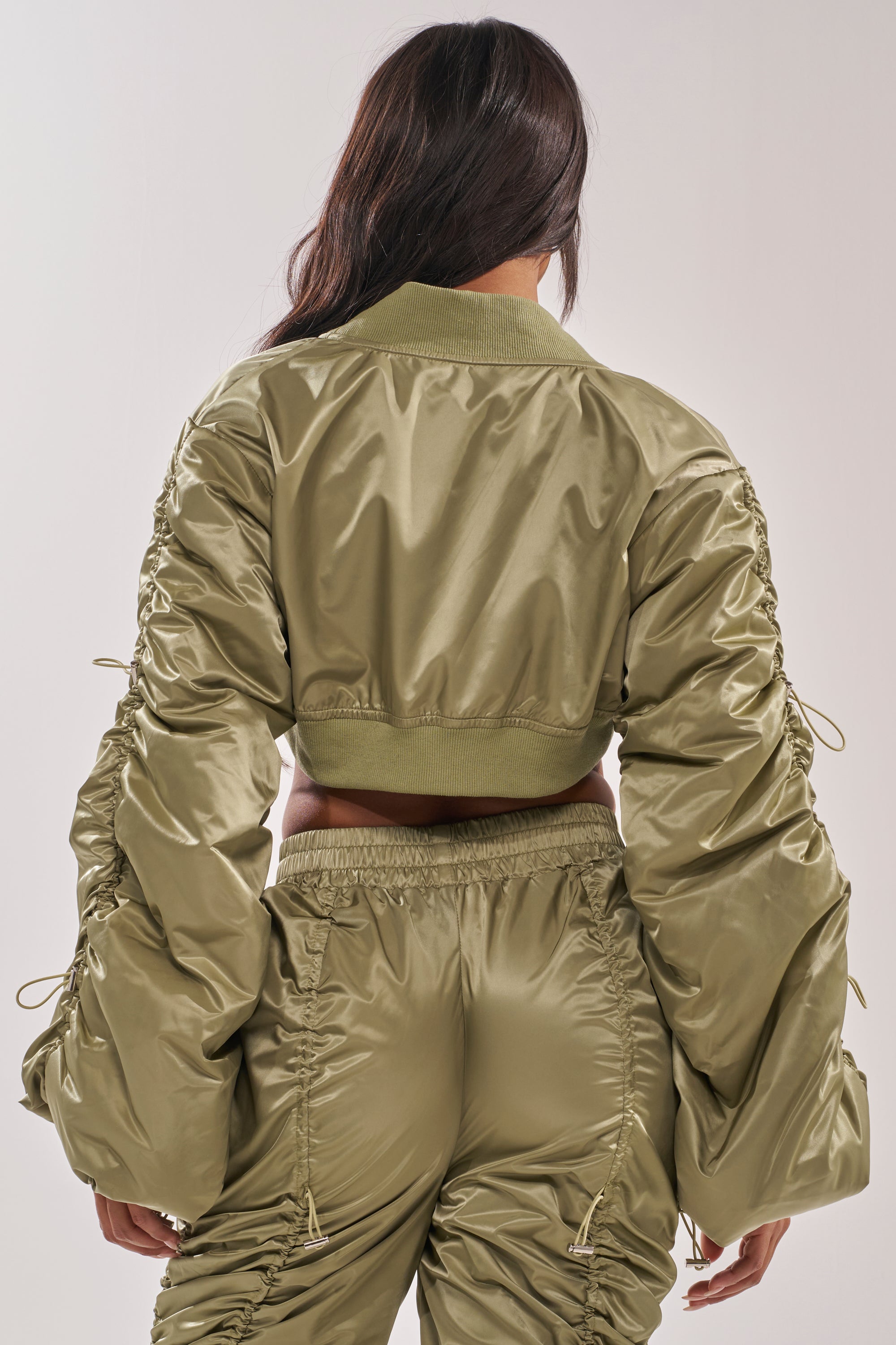 A woman is seen from the back wearing the NO TIME FOR GAMES BOLERO IN SAGE, a shiny, cropped bomber-style jacket with ruched sleeves, paired with matching glossy high-waisted pants. 3