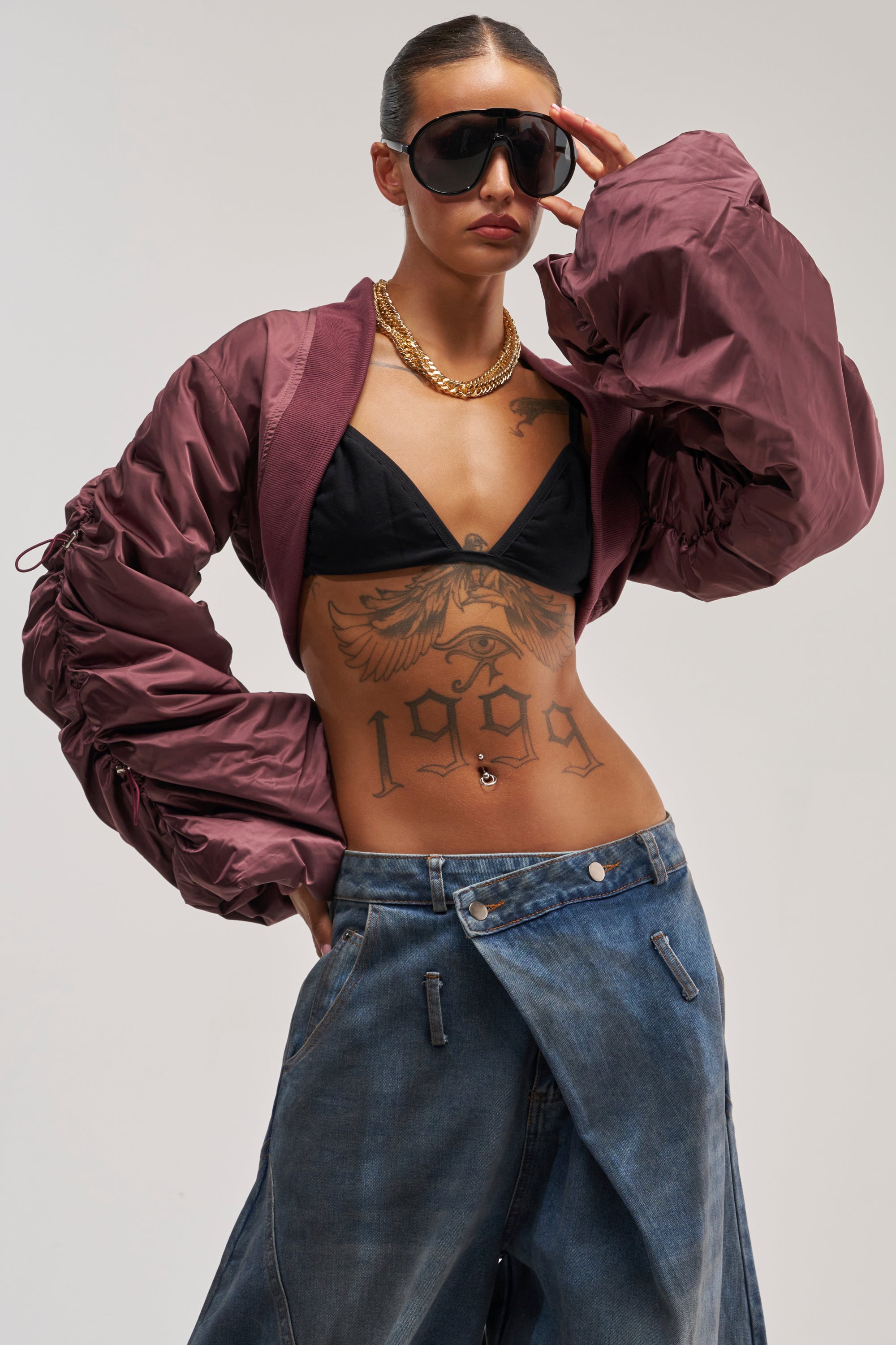 Wearing the NO TIME FOR GAMES RUCHED BOLERO, along with sunglasses, a gold chain, black bikini top, and baggy jeans, this person poses confidently, showcasing a large eagle and "1989" tattoo on their torso.