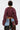 A person with short dark hair, wearing the NO TIME FOR GAMES RUCHED BOLERO in maroon and patchwork pants, stands facing away from the camera against a plain white background.