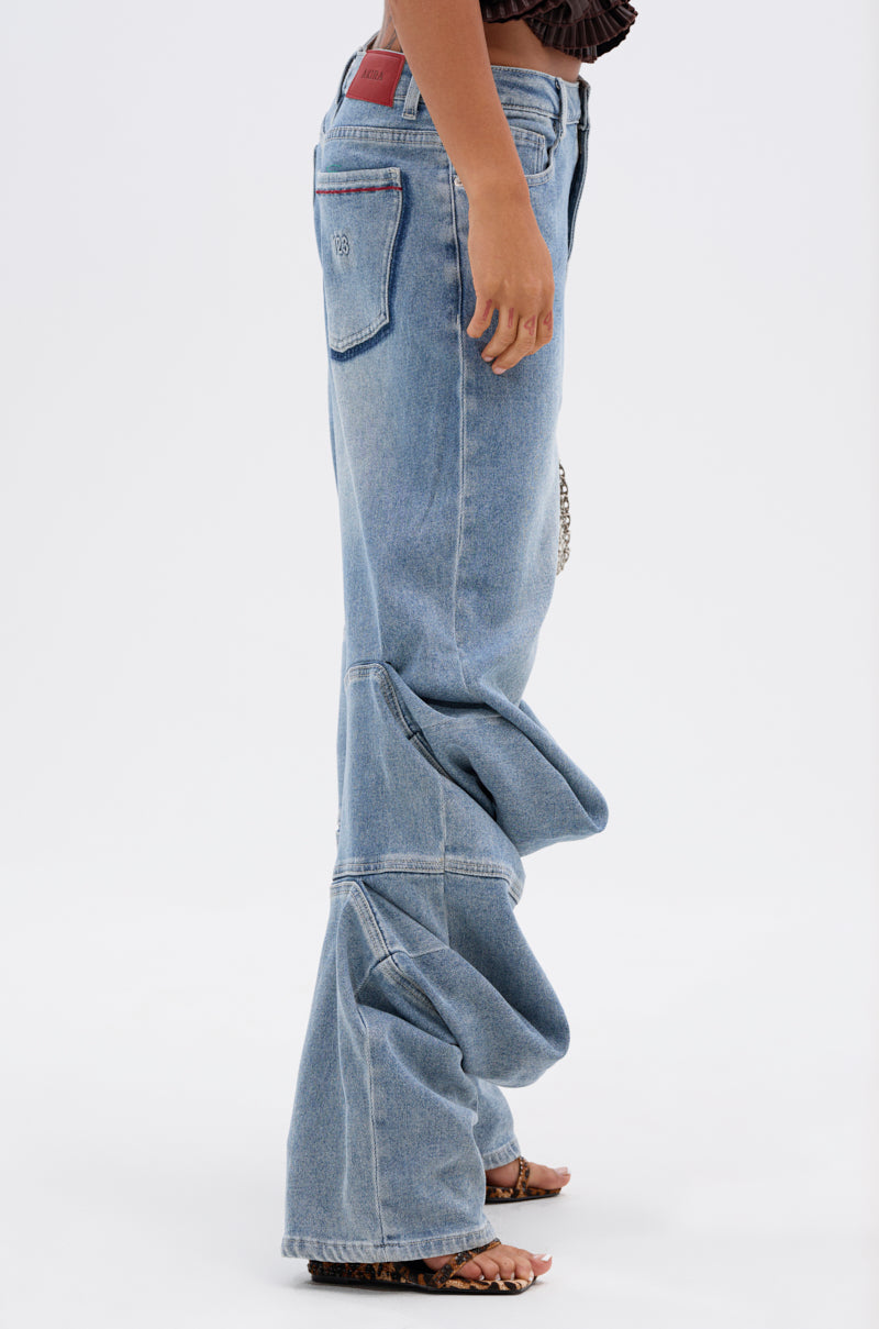 A person wears LAY LOW STACKED JEANS in light wash denim with exaggerated, bunched fabric at the ankles, paired with open-toe sandals. The background is plain white.