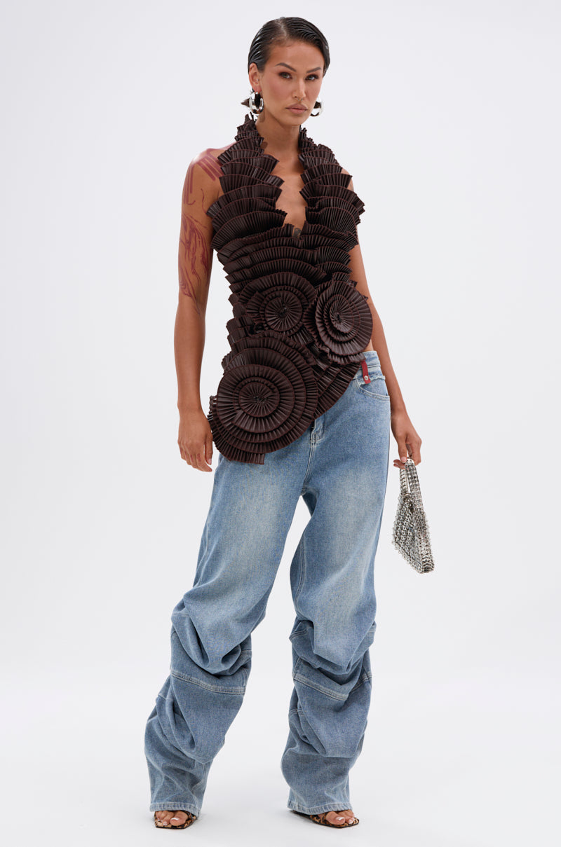 A person stands confidently in LAY LOW STACKED JEANS—light wash, baggy, mid-rise—paired with a sleeveless brown sculpted top with circular ruffle details, silver earrings, and a small silver handbag against a plain white background.