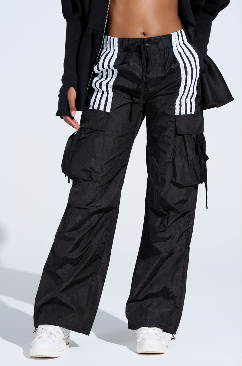 Wearing the JUMP AROUND TRACK PANT—black cargo pants with white vertical stripes at the waistband—paired with white sneakers and a black top, only the lower torso and legs are seen against a light background.