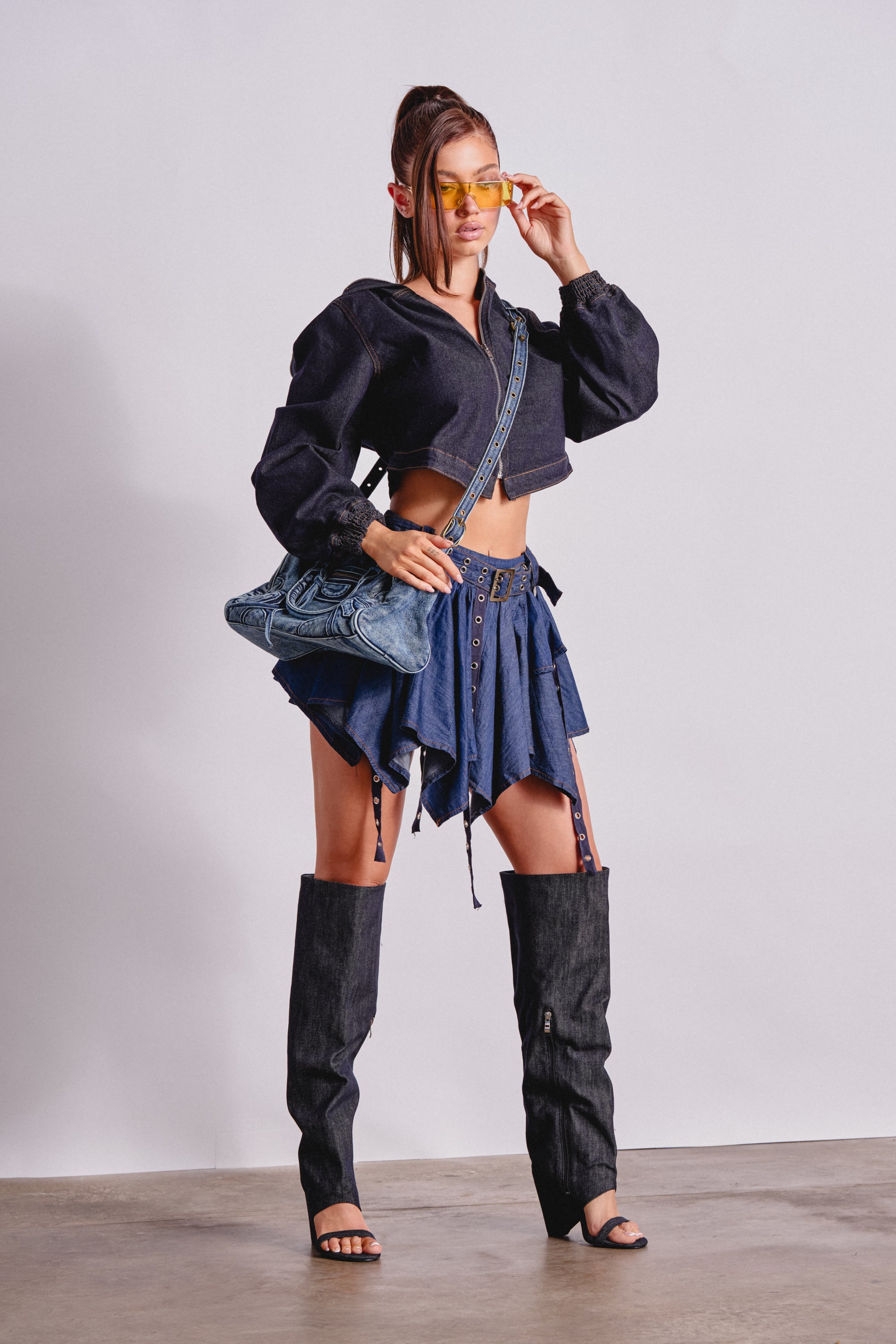 A woman poses against a plain background wearing a cropped black jacket, the AFTER HOURS MINI SKIRT IN MEDIUM BLUE DENIM, denim leg warmers, heeled sandals, yellow sunglasses, and a blue patterned crossbody bag. 0