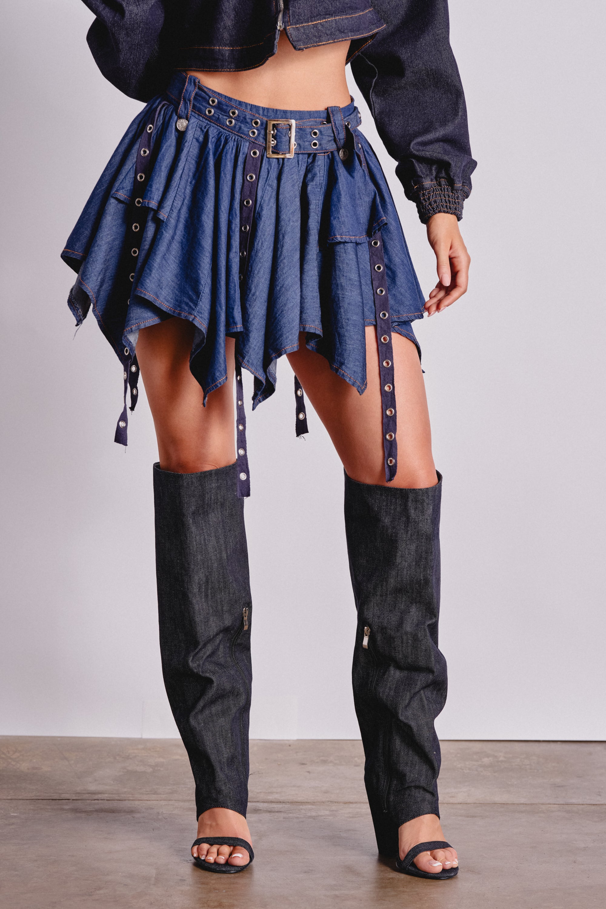 A person wears a dark denim cropped jacket, the AFTER HOURS MINI SKIRT IN MEDIUM BLUE DENIM with an asymmetrical handkerchief hem, and matching tall denim leg warmers over black heeled sandals—perfect for festivals. 2