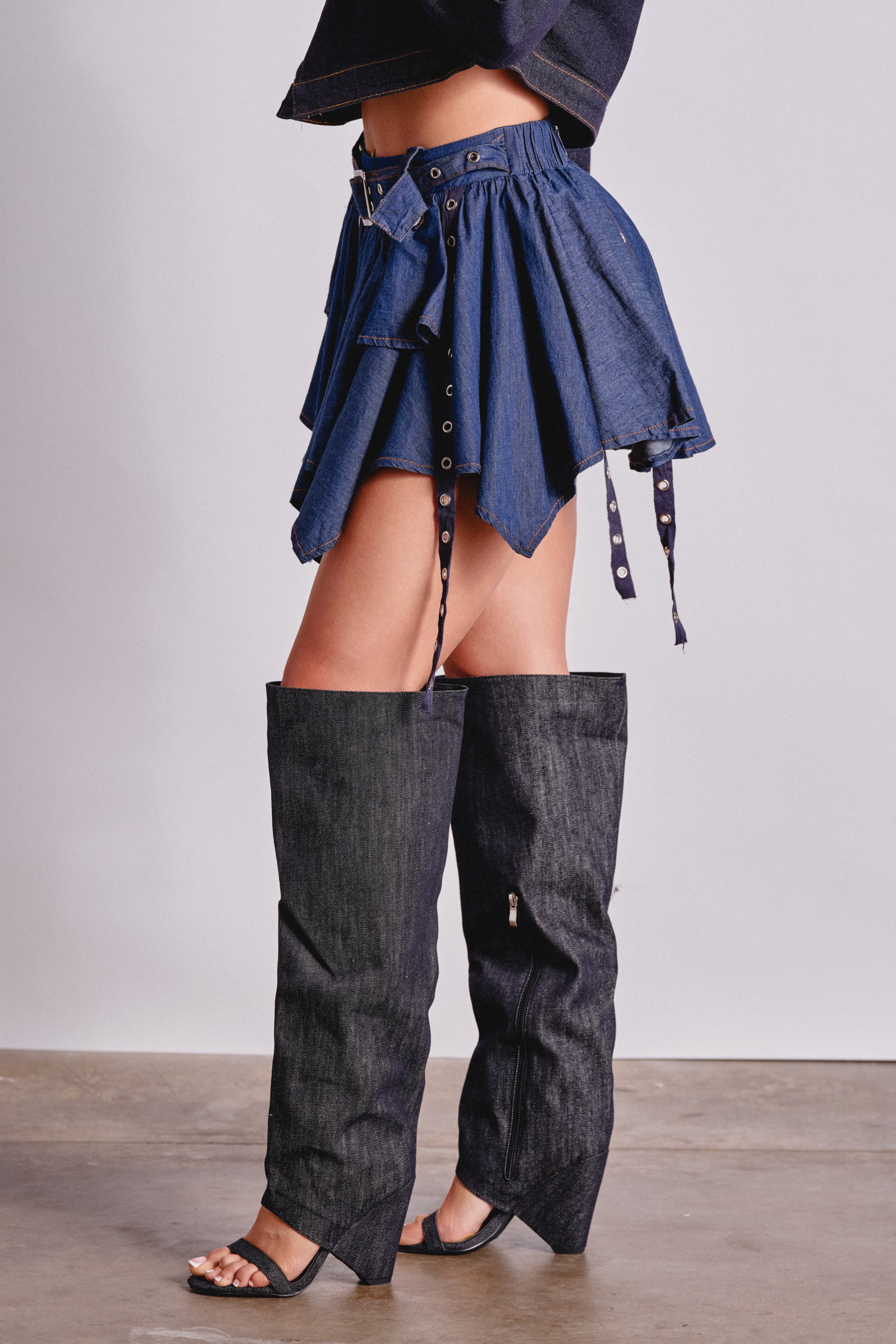 Wearing the AFTER HOURS MINI SKIRT IN MEDIUM BLUE DENIM with a handkerchief hem and oversized pant legs, paired with black heeled sandals, creates a standout festival outfit on a concrete floor. 3