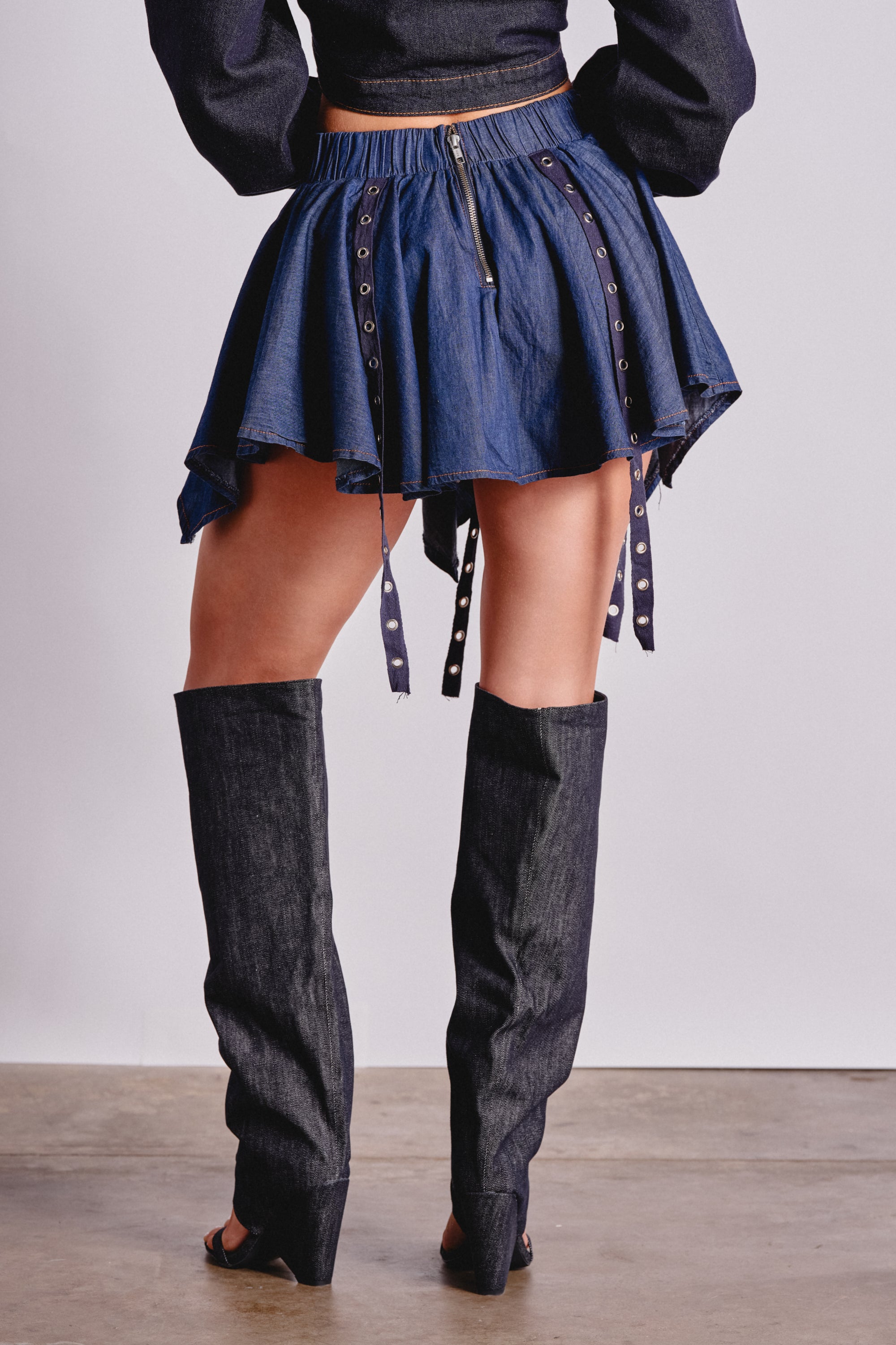 Wearing the AFTER HOURS MINI SKIRT IN MEDIUM BLUE DENIM with grommet and strap details and matching tall boots, a person stands on a concrete floor—creating a bold festival look against a plain, light-colored background. 4